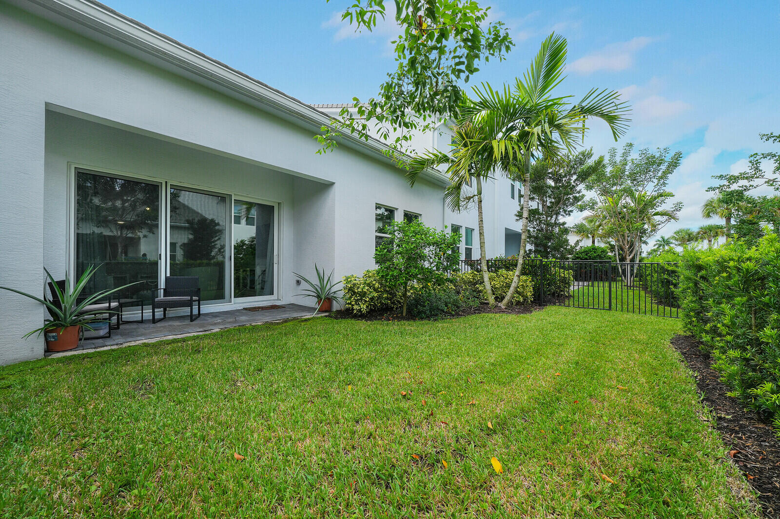 Rear exterior featuring a covered stone patio, sliding glass doors, manicured lawn, metal perimeter fencing, and mature palm landscaping