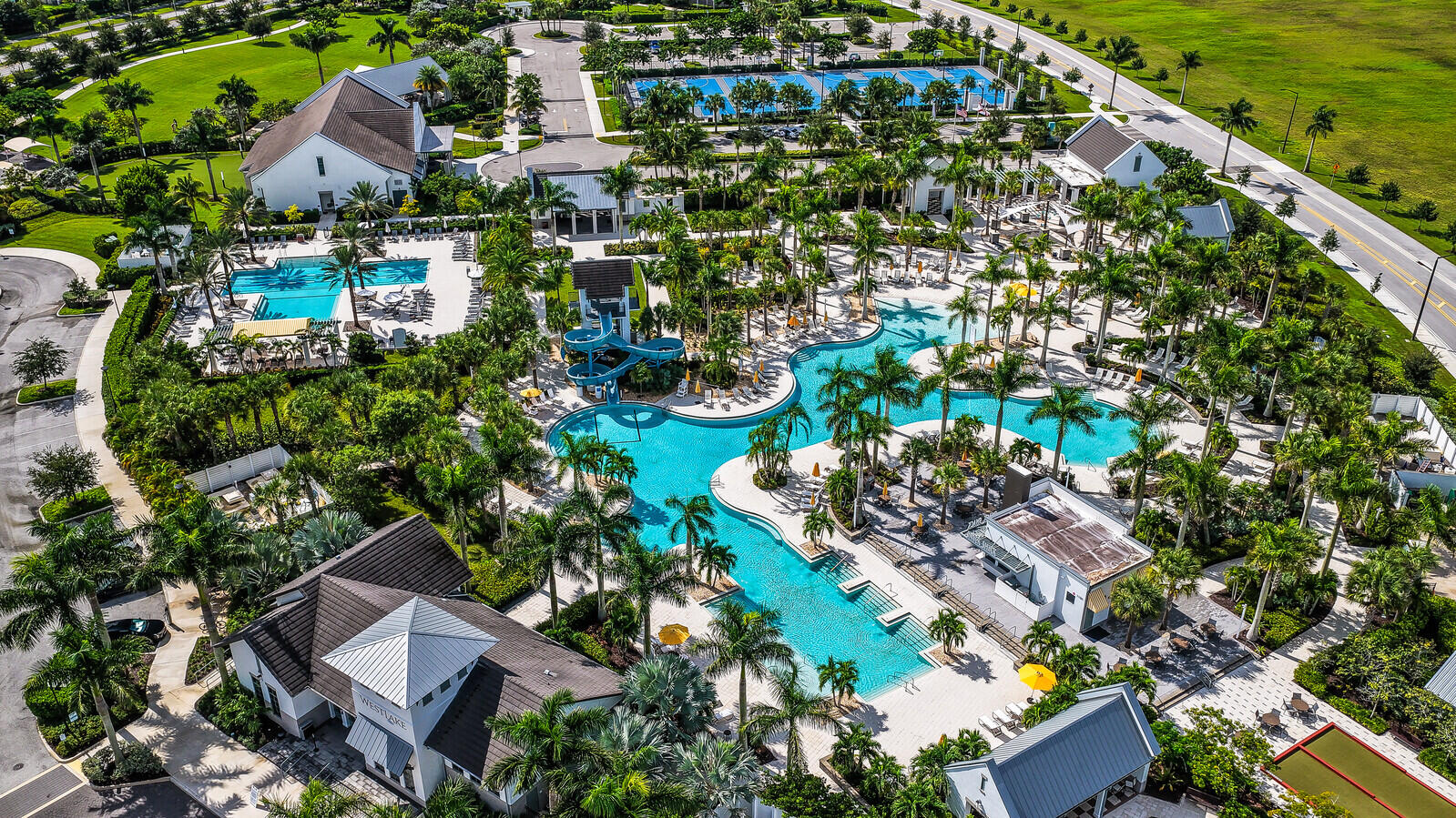 Aerial perspective of a community amenity complex featuring multiple swimming pools, water slides, paved terrace areas, and tennis courts