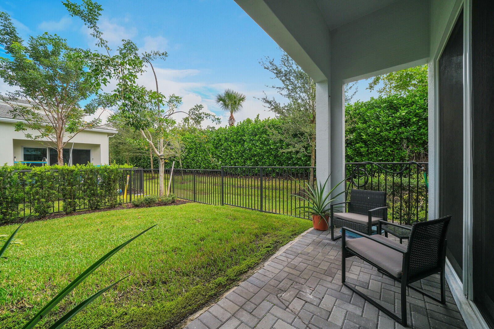 Enjoy serenity in your covered patio featuring paver flooring, enclosed backyard with manicured lawn, black metal fencing, and perimeter privacy hedges