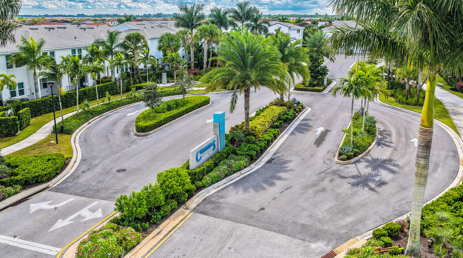 Gated entrance featuring landscaped medians, mature palm trees, paved roadways, and a prominent community monument sign