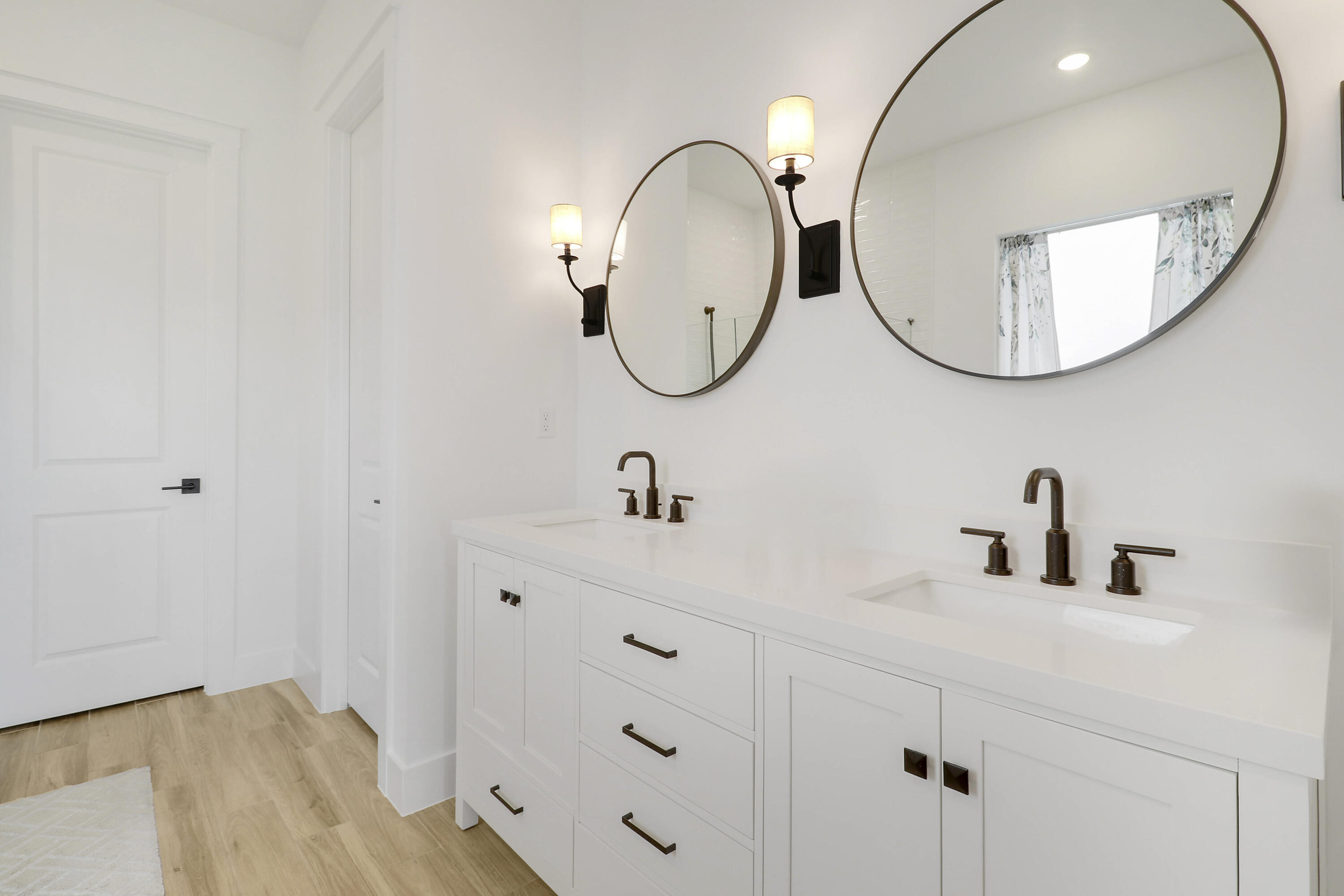 Dual-sink vanity featuring a white composite countertop, undermount sinks, matte black widespread faucets, and shaker-style cabinetry with matte black hardware, complemented by round-frame mirrors and wall-mounted sconces