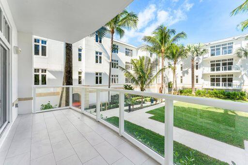 Covered balcony featuring large-format tile flooring and a glass panel railing, overlooking a landscaped courtyard with a manicured lawn, paved walkways, and palm trees Covered balcony featuring large-format tile flooring and a glass panel railing, overlooking a landscaped courtyard with a manicured lawn, paved walkways, and palm trees
