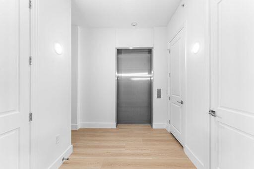 Interior hallway featuring wood-finish flooring, brushed metal elevator doors, two-panel interior doors with lever-style handles, and circular wall-mounted lighting