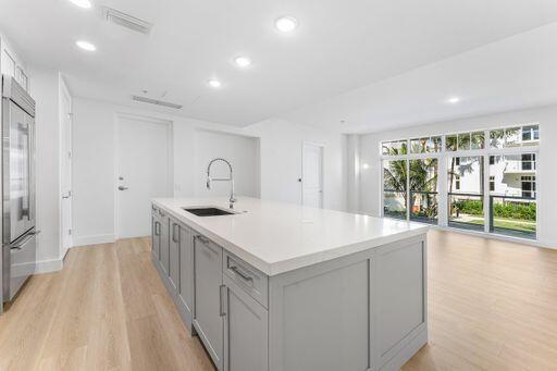 Kitchen featuring a large central island with light-gray shaker cabinetry, a white countertop, an undermount sink, and a gooseneck faucet, set against wood-finish flooring and expansive multi-panel glass doors Kitchen featuring a large central island with light-gray shaker cabinetry, a white countertop, an undermount sink, and a gooseneck faucet, set against wood-finish flooring and expansive multi-panel glass doors