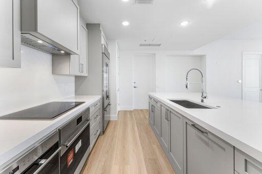 Kitchen featuring white countertops, light gray shaker-style cabinetry, stainless steel appliances, a center island with an integrated sink, and light wood-finish flooring Kitchen featuring white countertops, light gray shaker-style cabinetry, stainless steel appliances, a center island with an integrated sink, and light wood-finish flooring