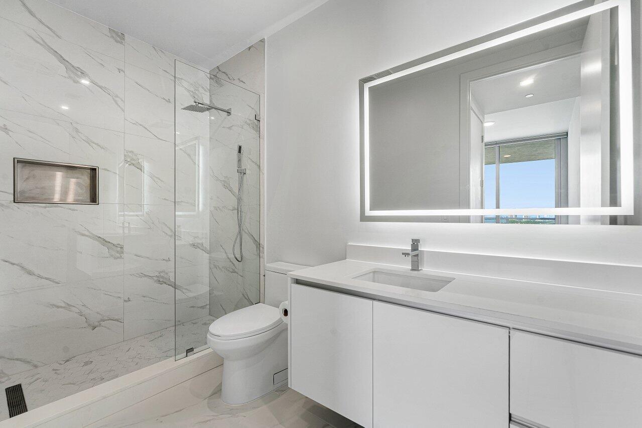 Bathroom featuring floor-to-ceiling stone-look tiled shower with integrated niche and glass enclosure, illuminated vanity mirror, and flat-panel cabinetry