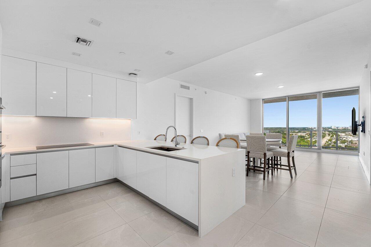 Modern kitchen featuring flat-panel white cabinetry, quartz countertops, a central island with an integrated sink, recessed lighting, and expansive floor-to-ceiling windows