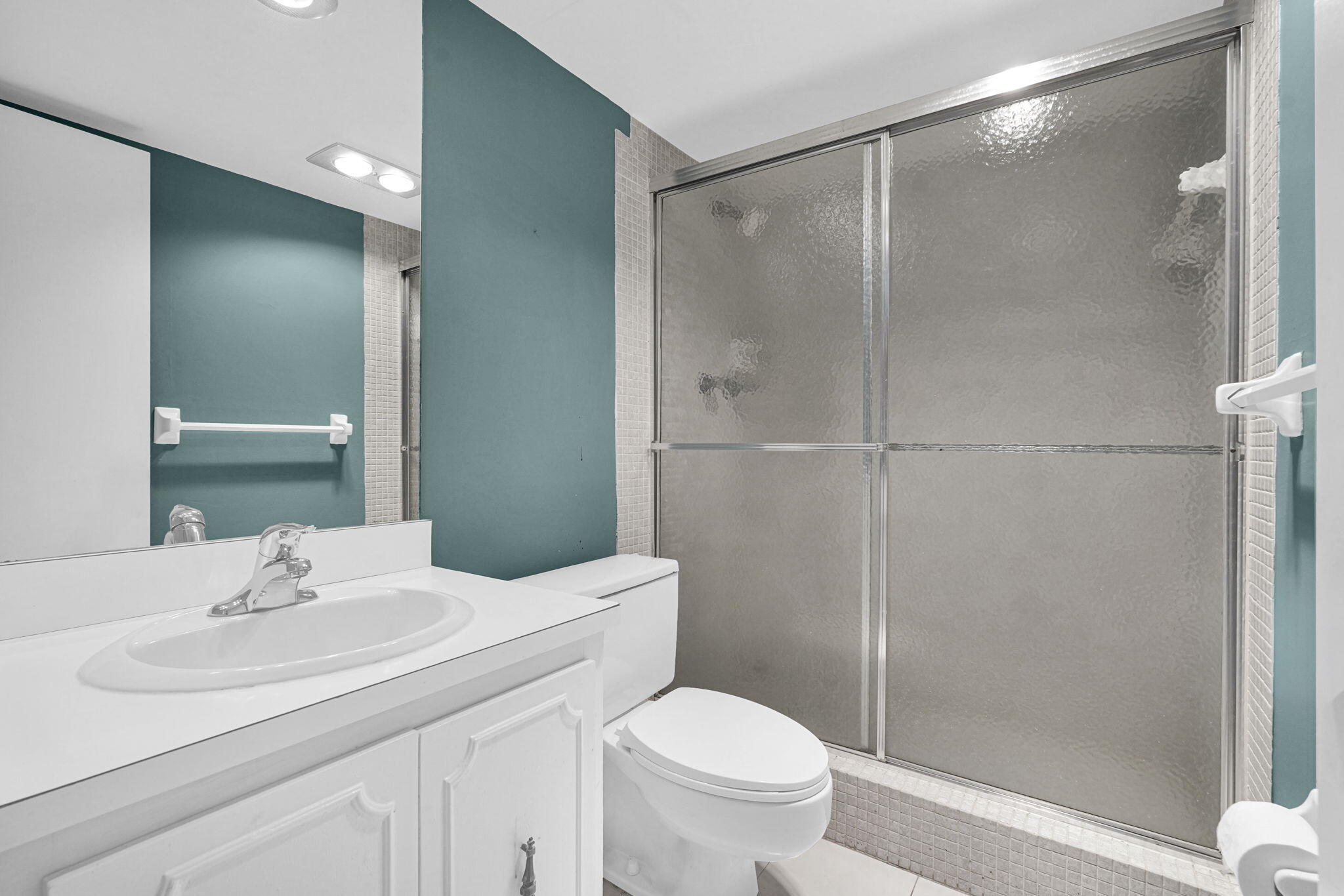 Bathroom featuring a white vanity cabinet with a solid surface countertop, integrated oval sink, chrome faucet, wall-to-wall mirror, and a walk-in shower with a sliding, textured-glass enclosure