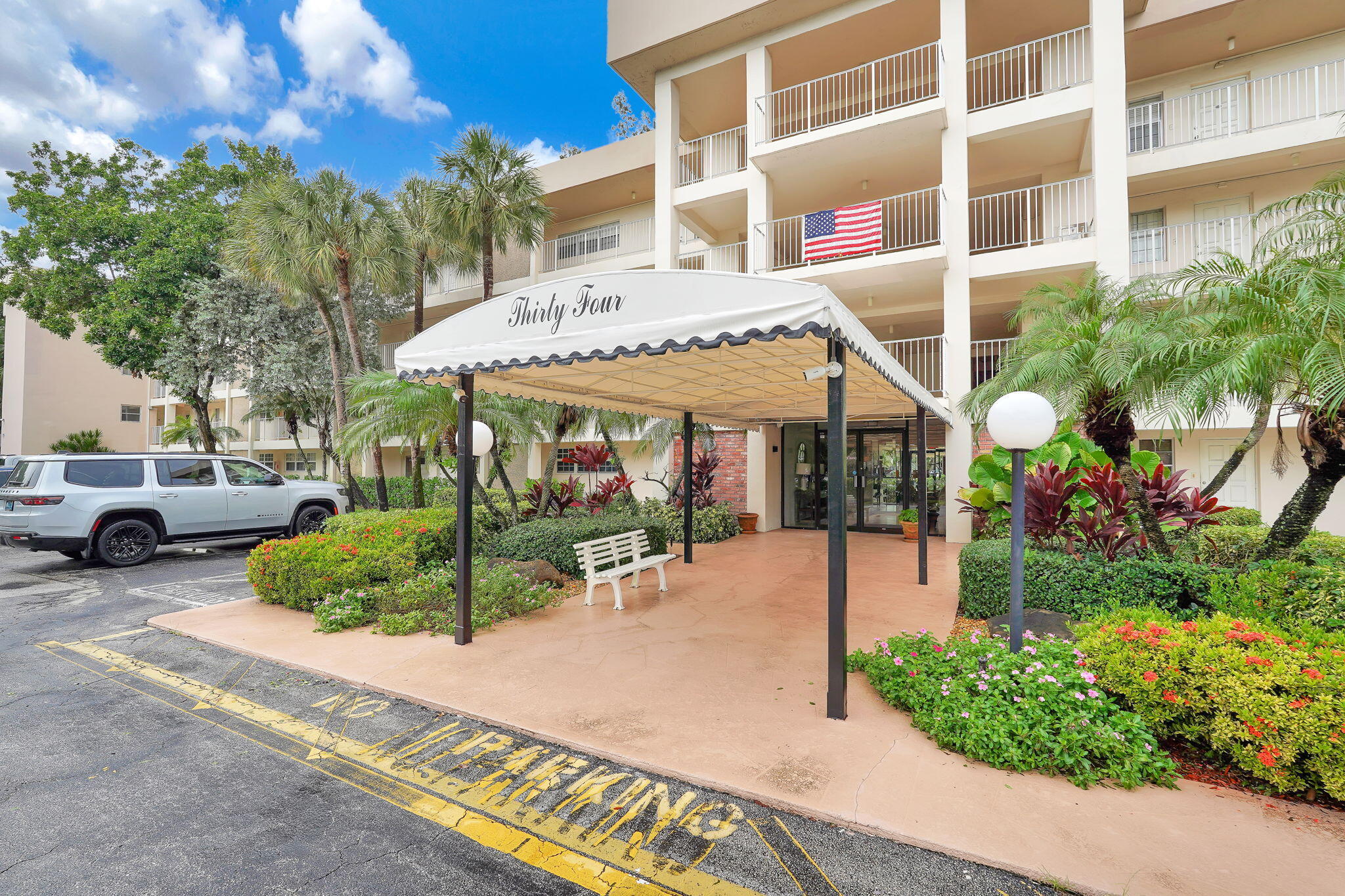Building exterior featuring a covered porte-cochre, multi-level balconies with railings, and manicured landscaping with mature palm trees