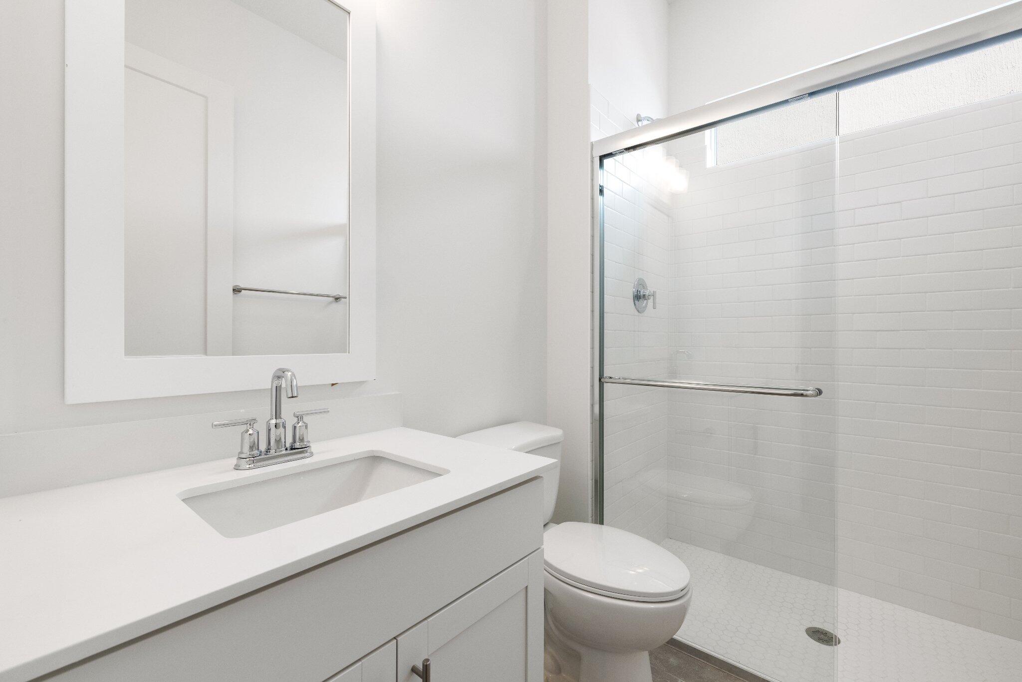 White vanity with a solid-surface countertop and undermount sink, framed rectangular wall mirror, and walk-in glass-enclosed shower featuring white subway-style wall tile