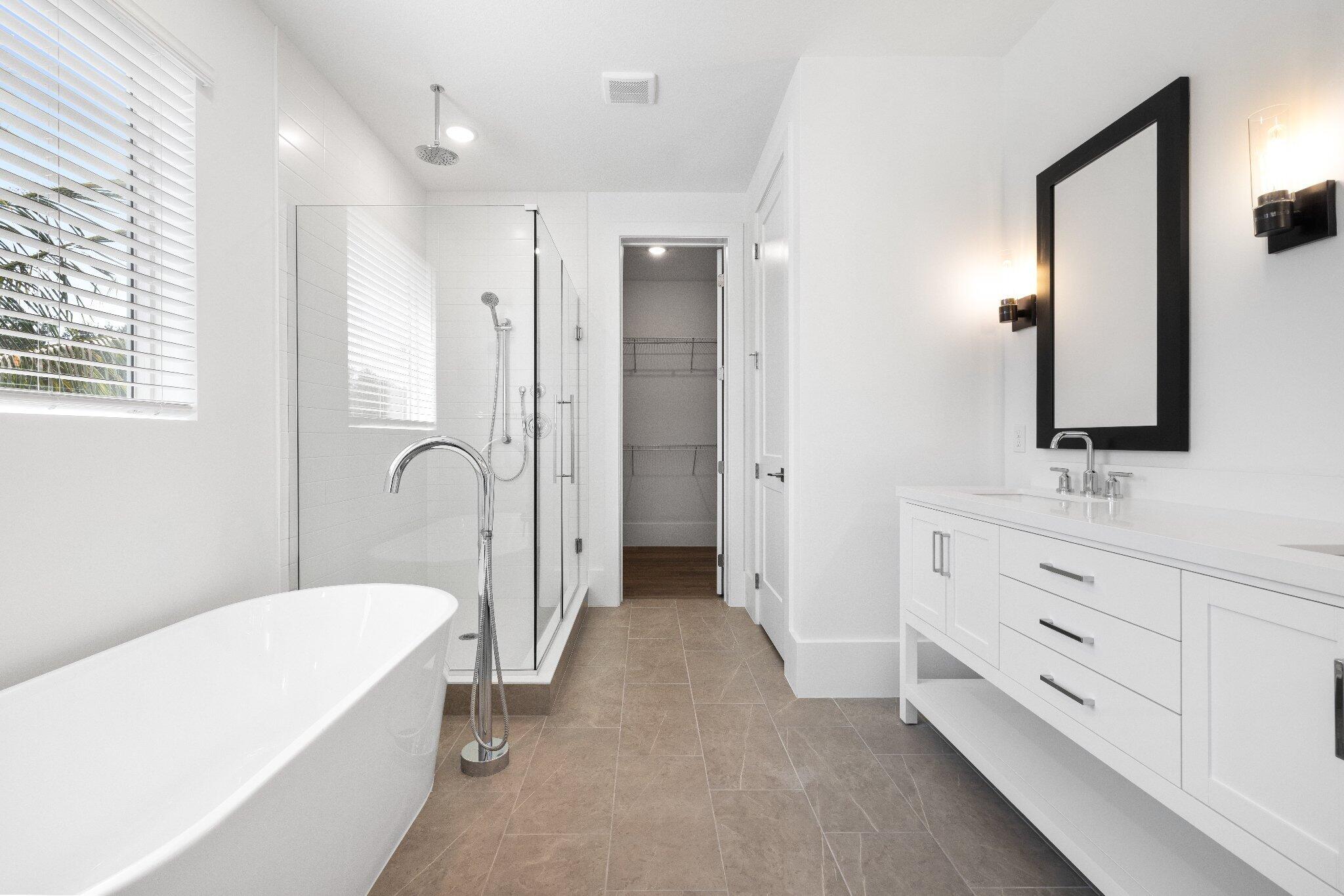 Freestanding soaking tub with floor-mounted faucet, walk-in glass-enclosed shower, white dual-sink vanity with countertop, framed vanity mirrors, and tile flooring