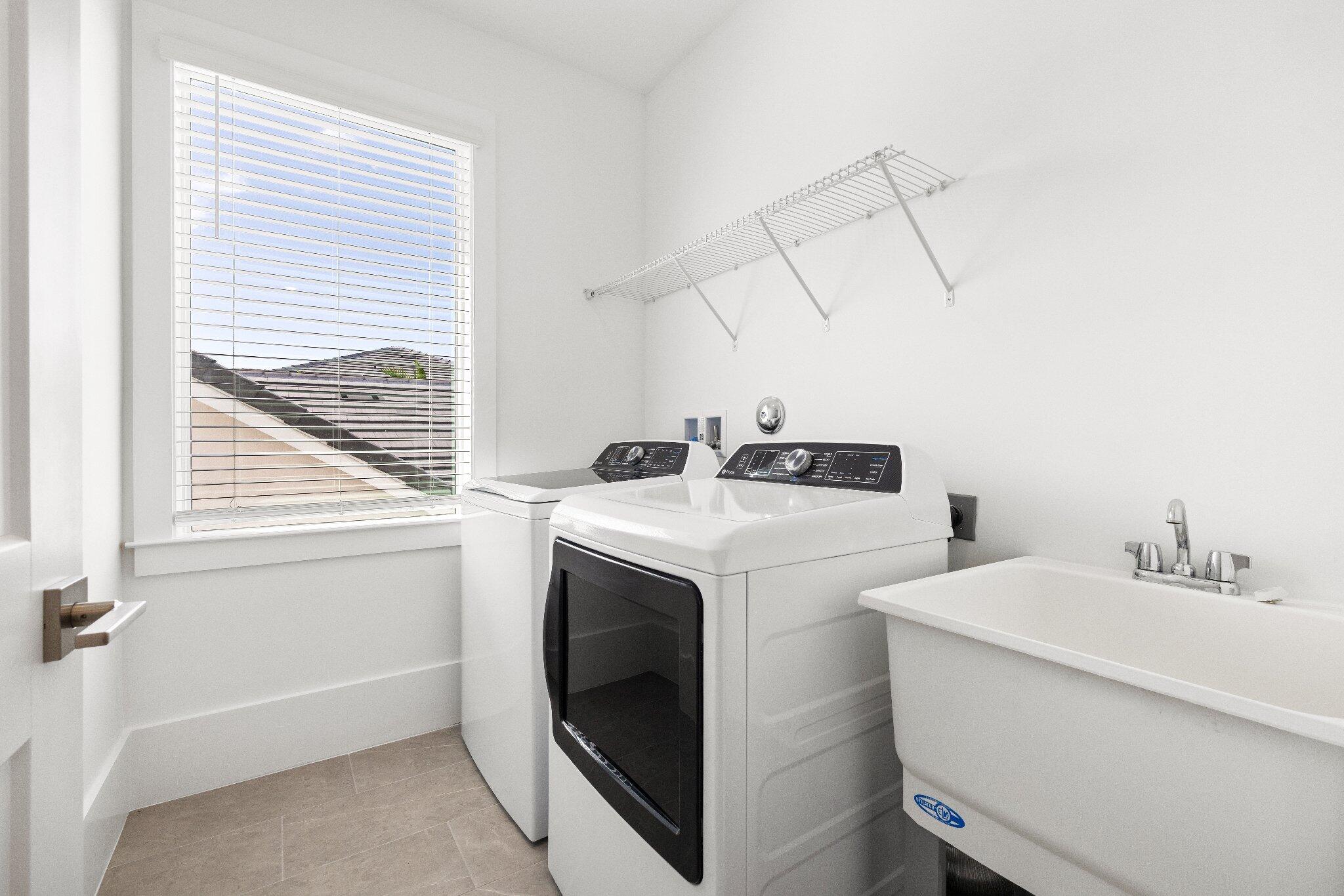 Laundry room featuring a deep utility sink, wall-mounted wire shelving, large window with horizontal blinds, and tile flooring