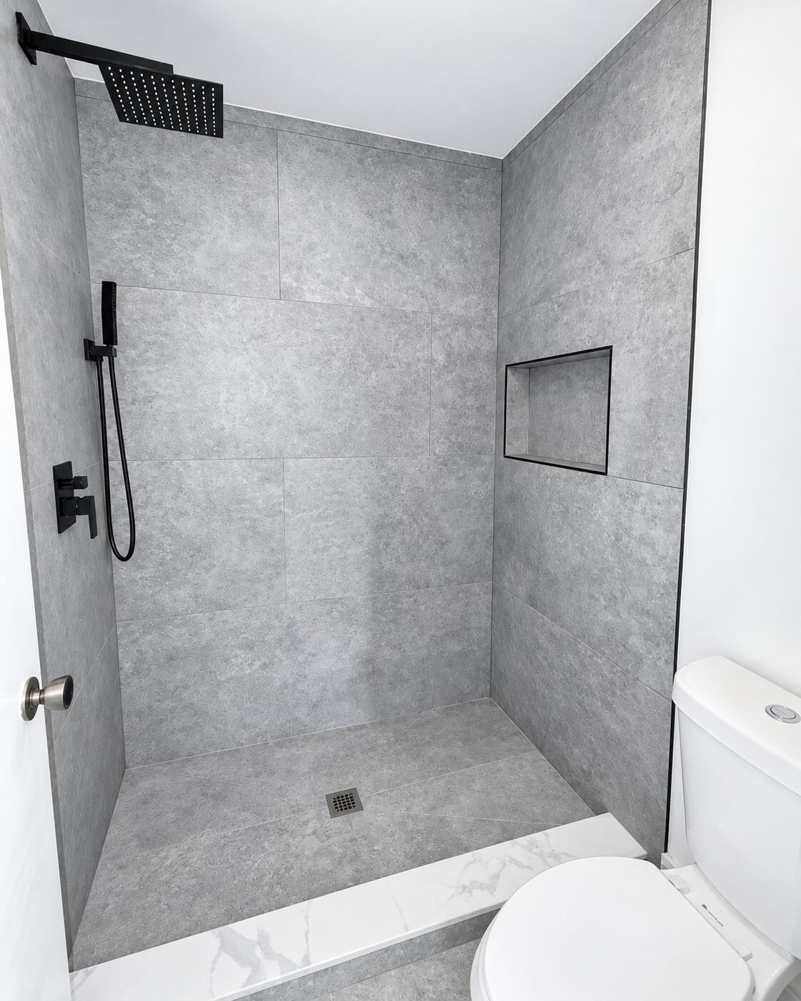 Walk-in shower featuring large-format grey tile, a matte black rainfall showerhead, a matching handheld wand, and a built-in recessed wall niche