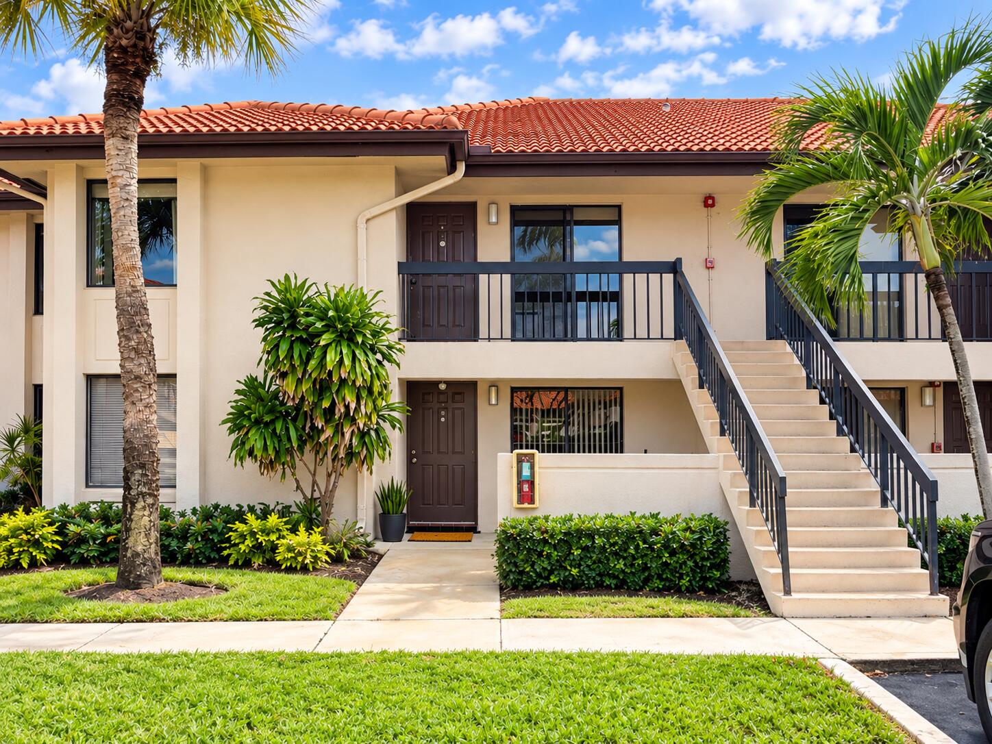 Stucco exterior featuring a terracotta tile roof, second-level railed balcony, concrete staircase, and tropical landscaping