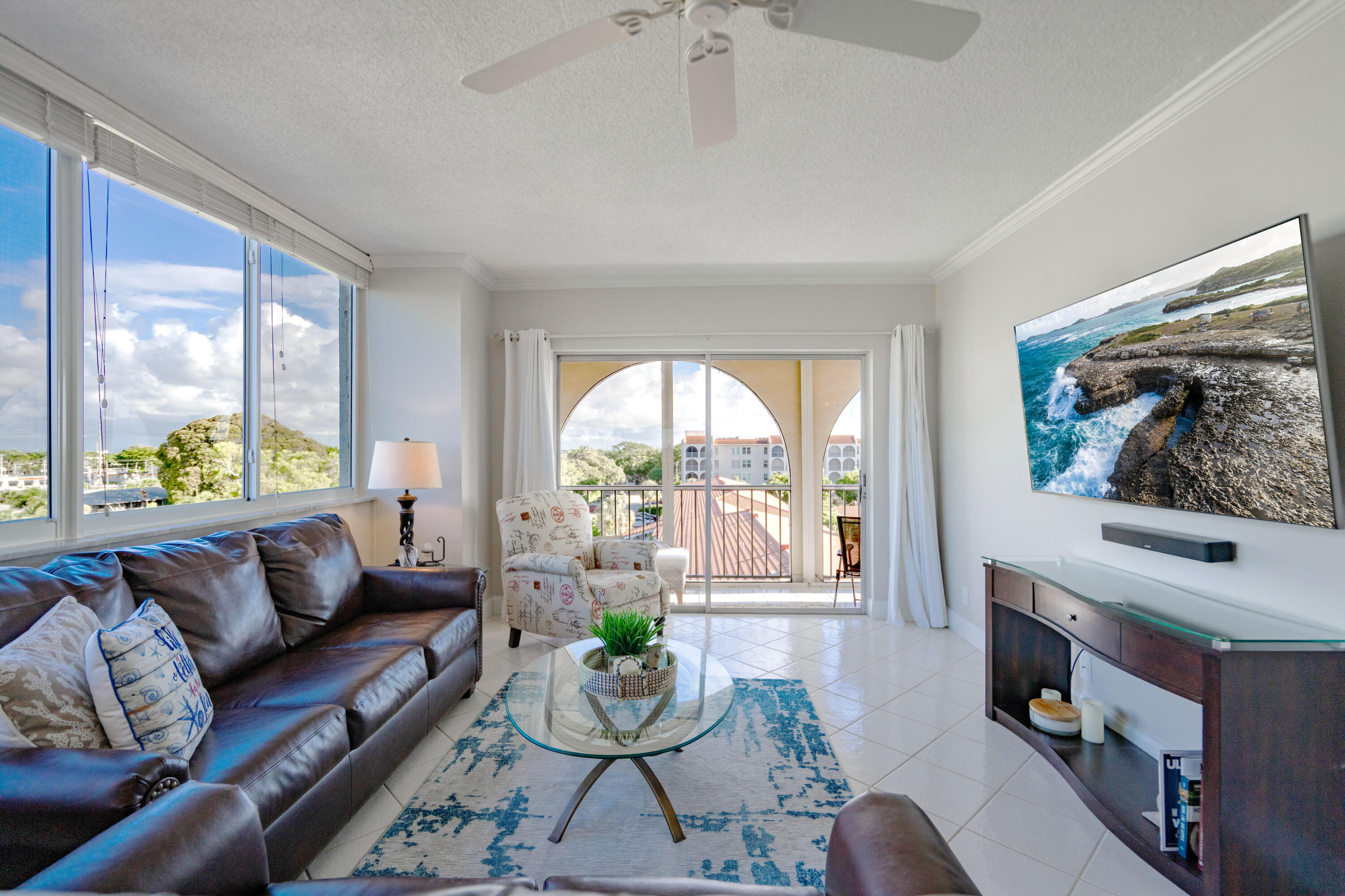 Living area featuring ceramic tile flooring, expansive windows, crown molding, a ceiling fan, and sliding glass doors with a decorative arched balcony enclosure