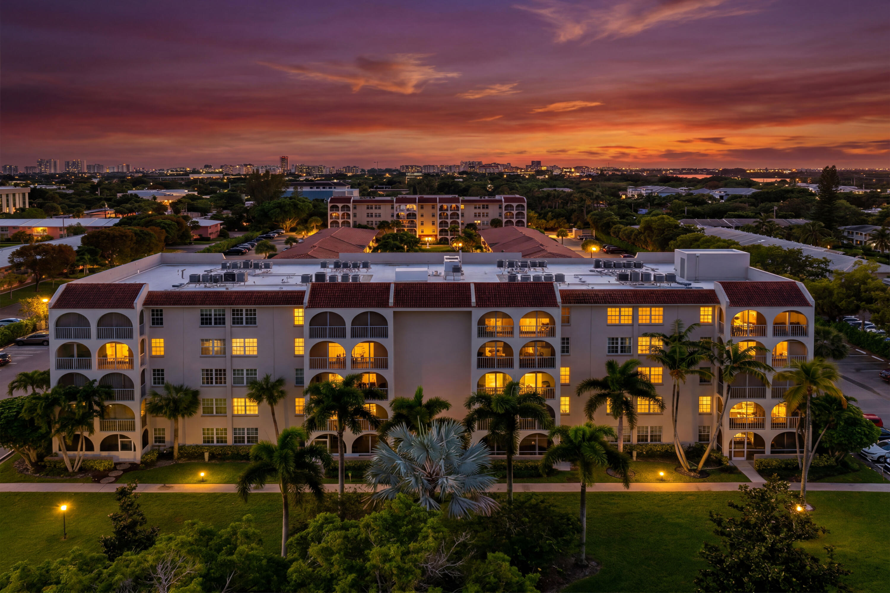 Multi-story residential building featuring arched balcony enclosures, a decorative tile roof, and manicured landscaping with mature palm trees