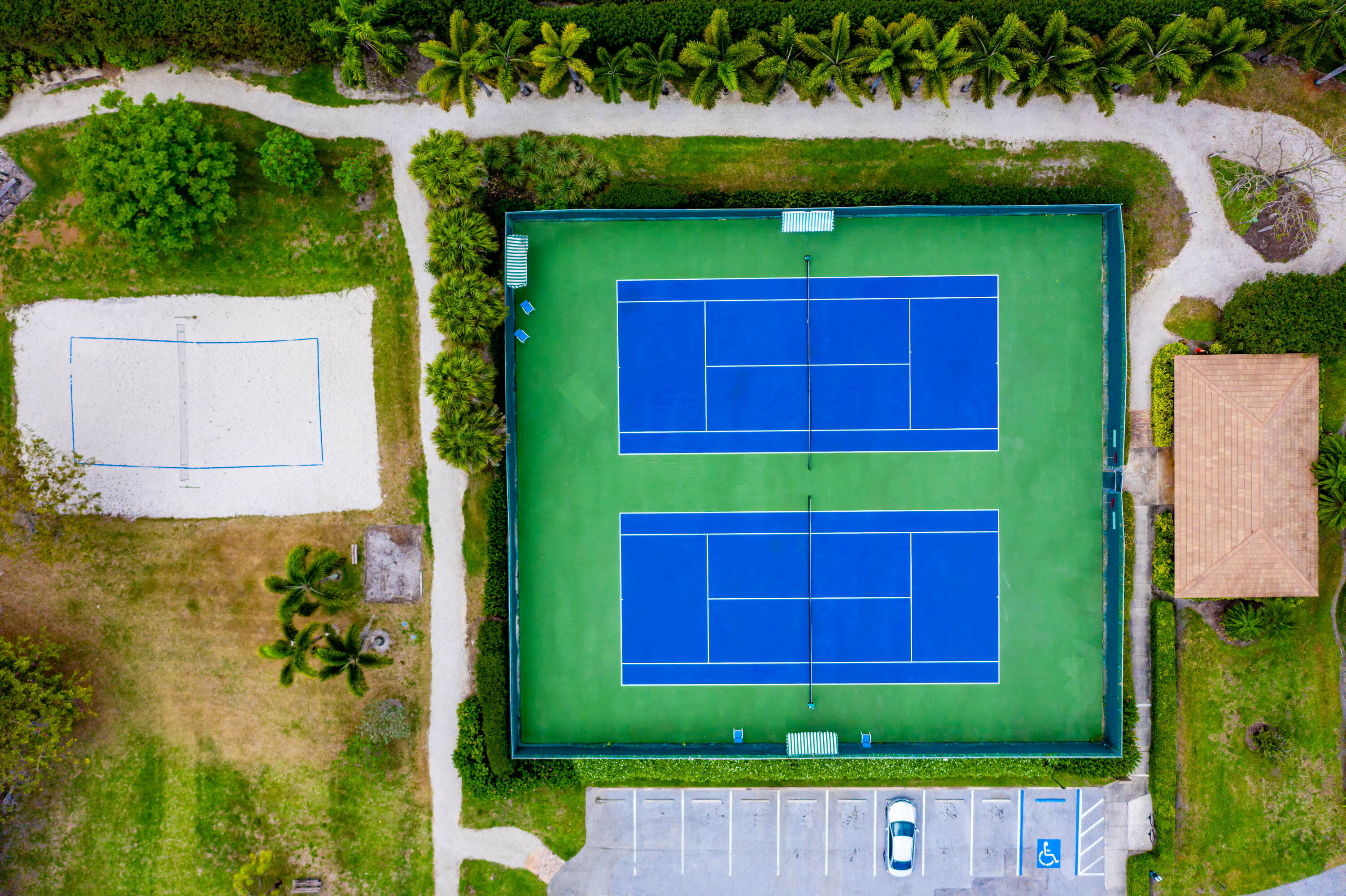 Dual tennis courts with fencing, adjacent sand court, and paved parking area surrounded by mature landscaping