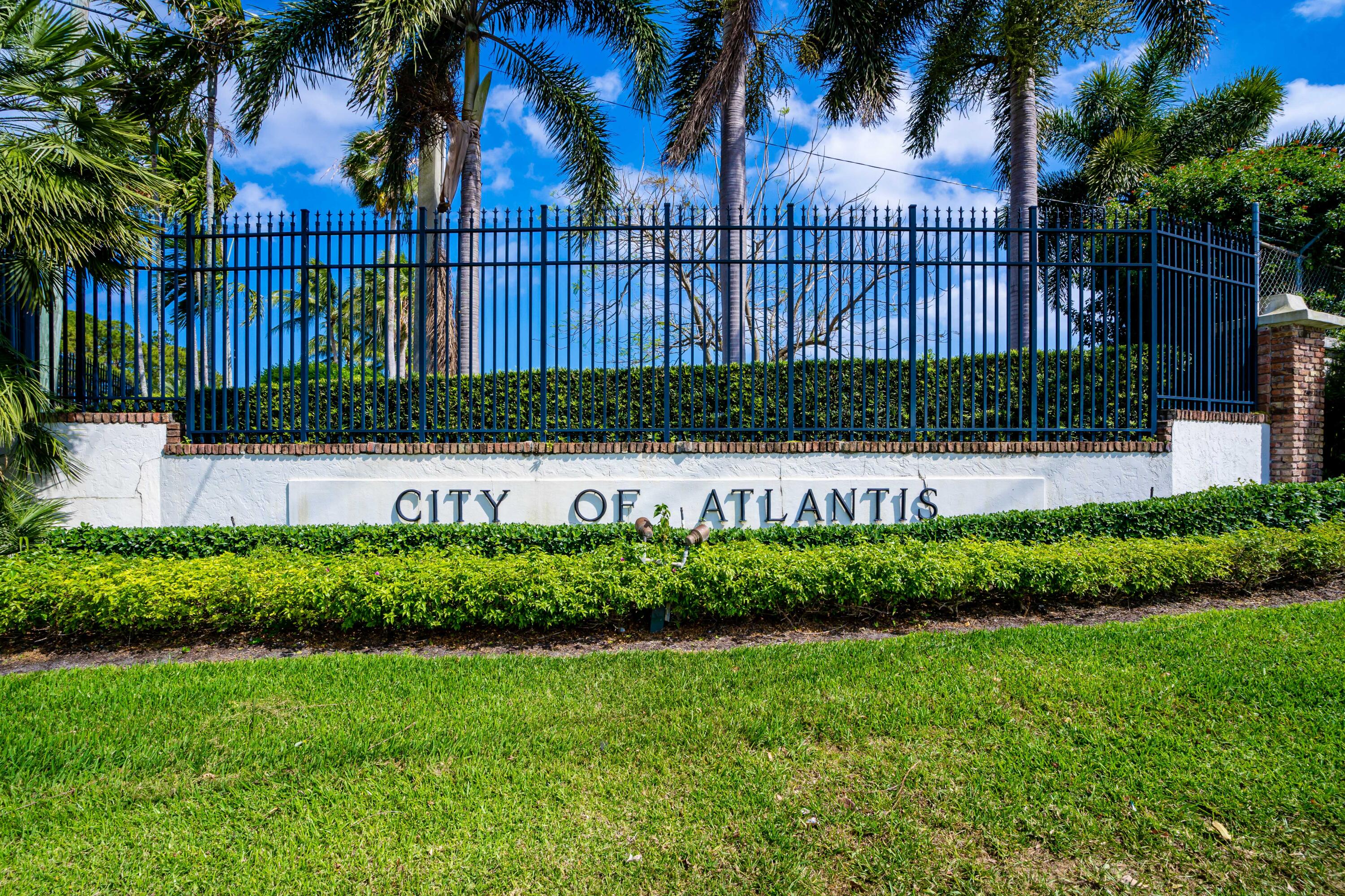 Community entrance featuring a concrete masonry wall, black metal fencing, brick pillar accents, and professionally manicured landscaping