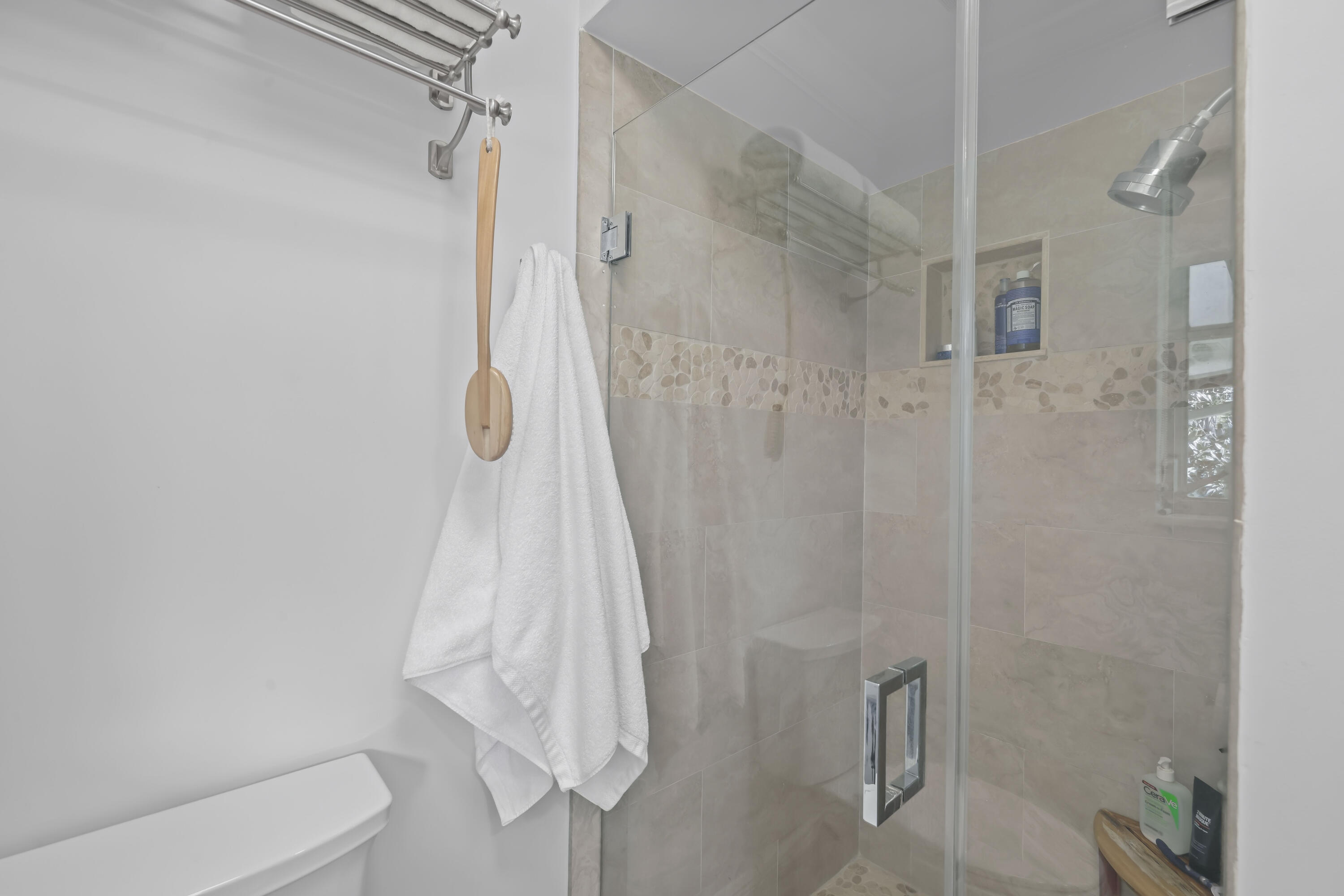 Shower featuring neutral stone-finish tile surround with a decorative mosaic inlay, a glass door enclosure, a built-in recessed shelf, and a polished metal shower head