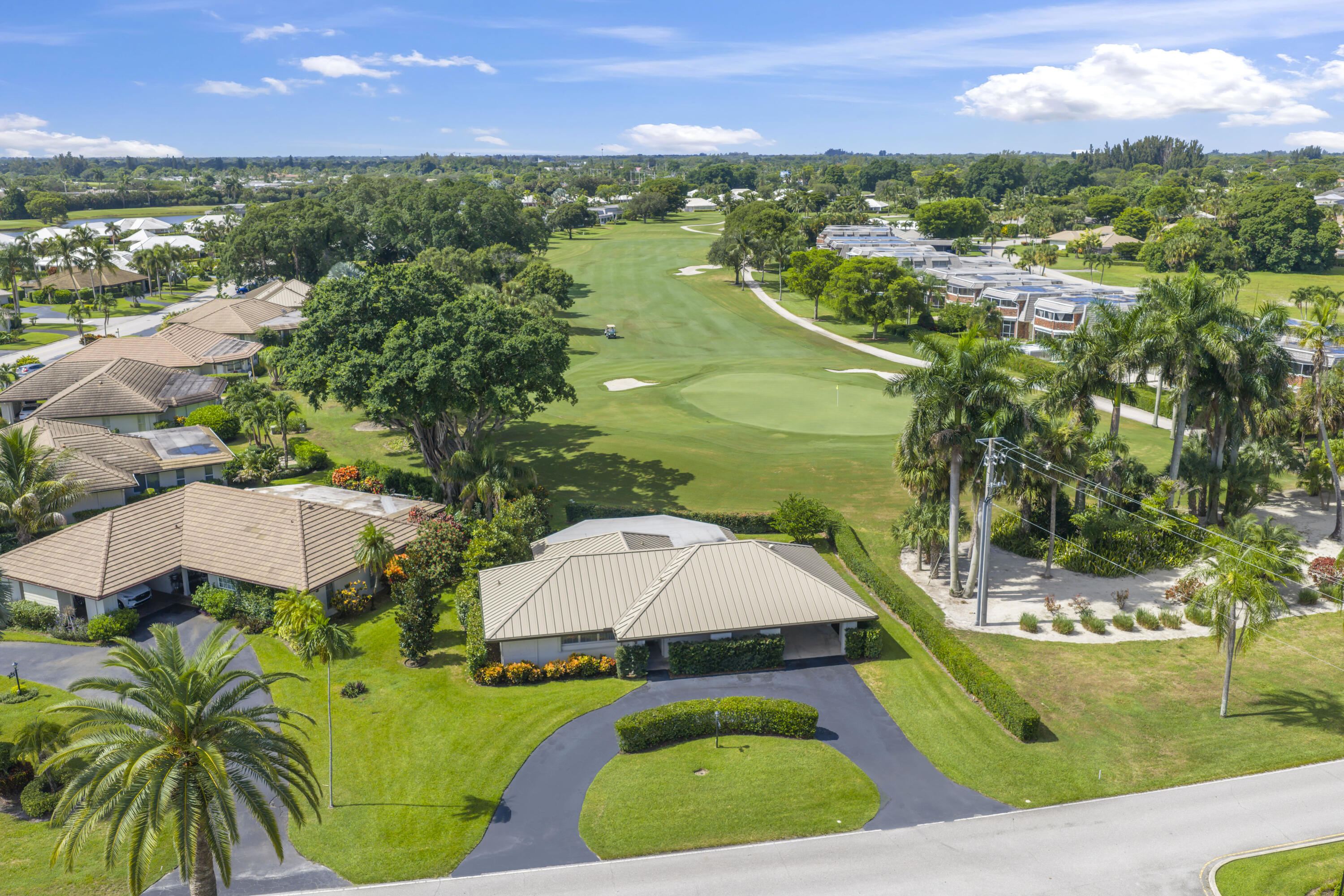 Aerial perspective featuring a single-story residence with a multi-hipped metal roof, curved asphalt driveway, and manicured lawn, situated on a golf course fairway
