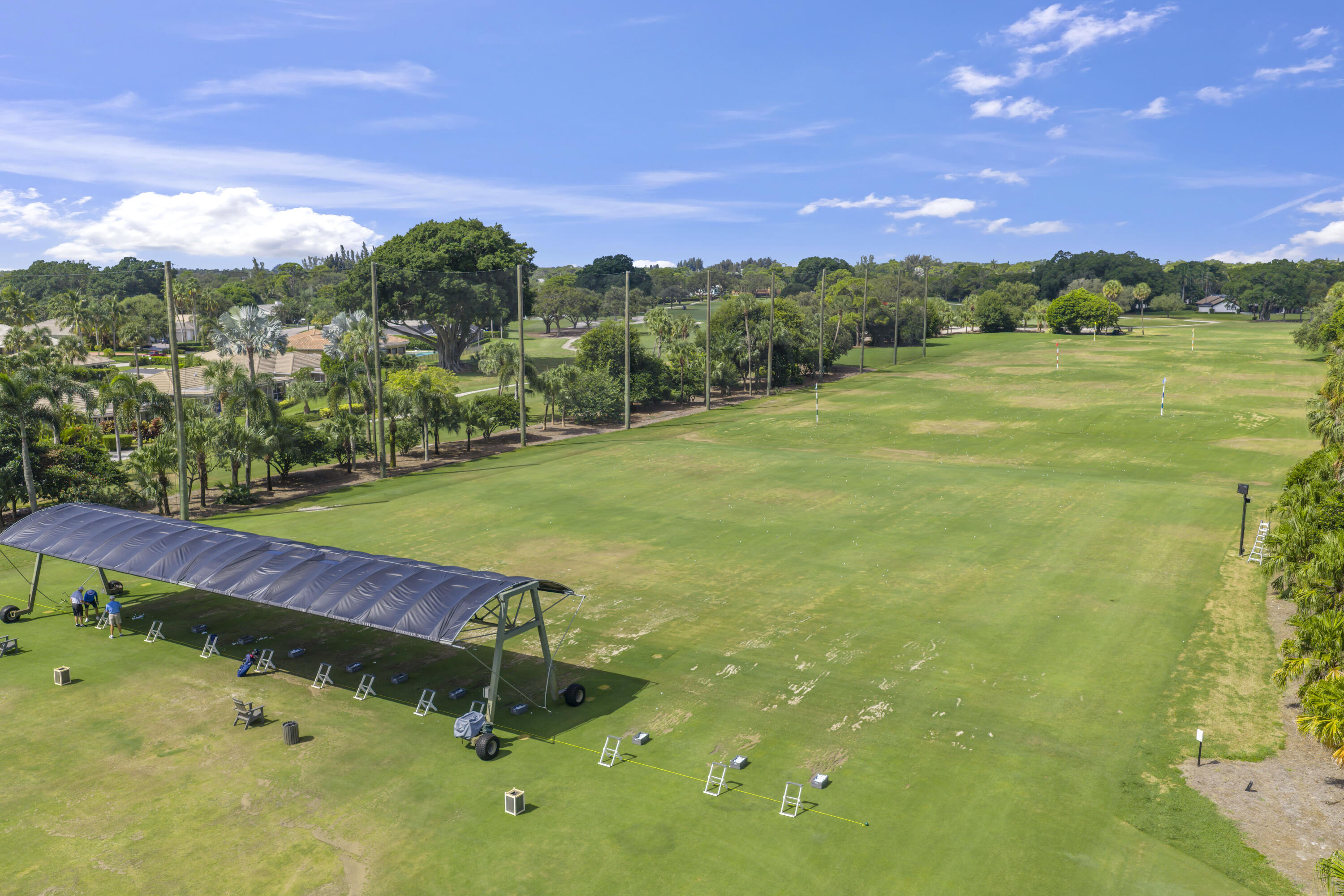 Expansive golf driving range featuring a covered hitting structure, multiple hitting bays, maintained turf, and perimeter landscaping with mature trees