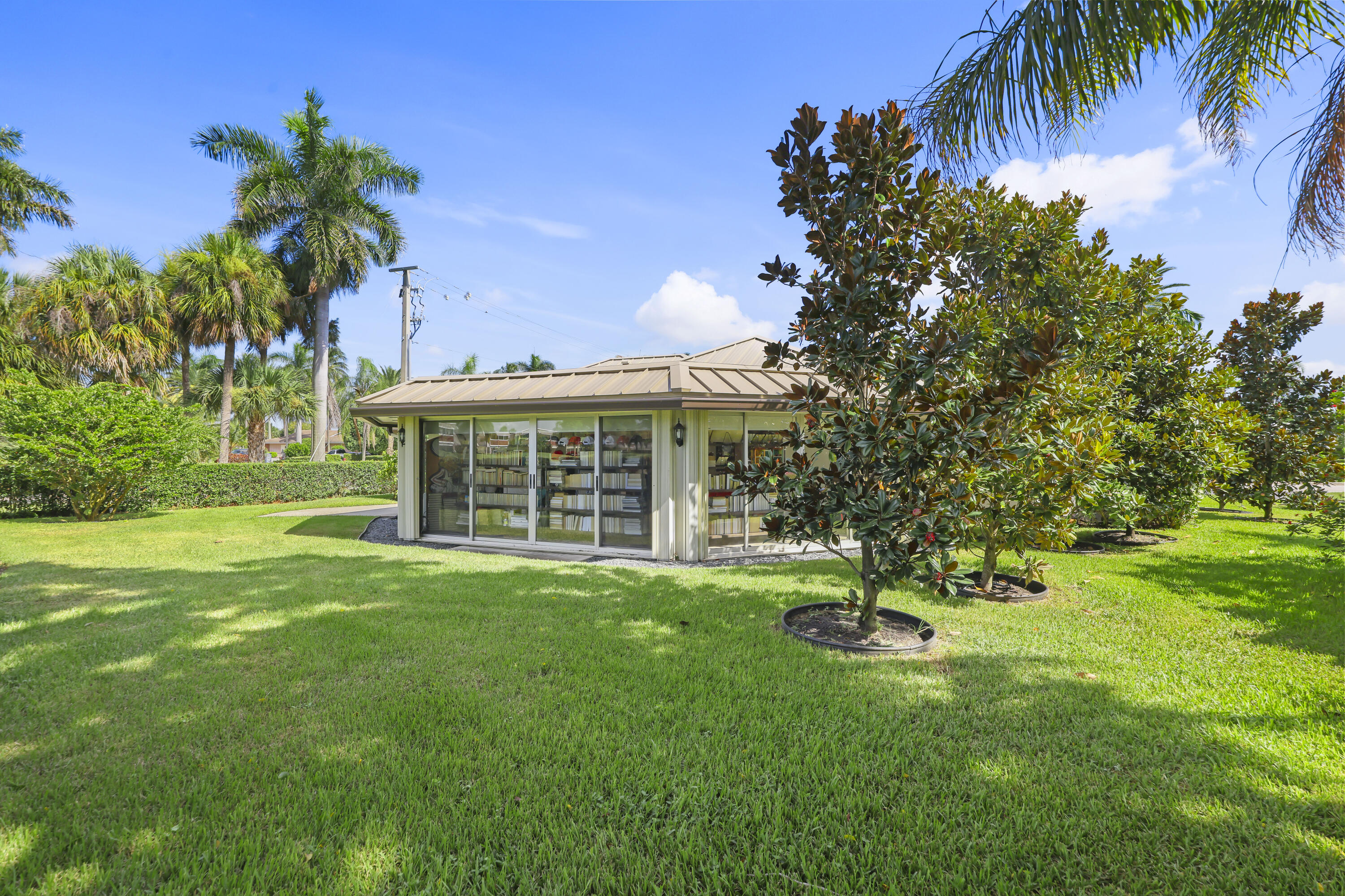Standalone structure featuring a metal hip roof, sliding glass wall panels, exterior wall-mounted lighting, and expansive landscaped grounds with mature trees