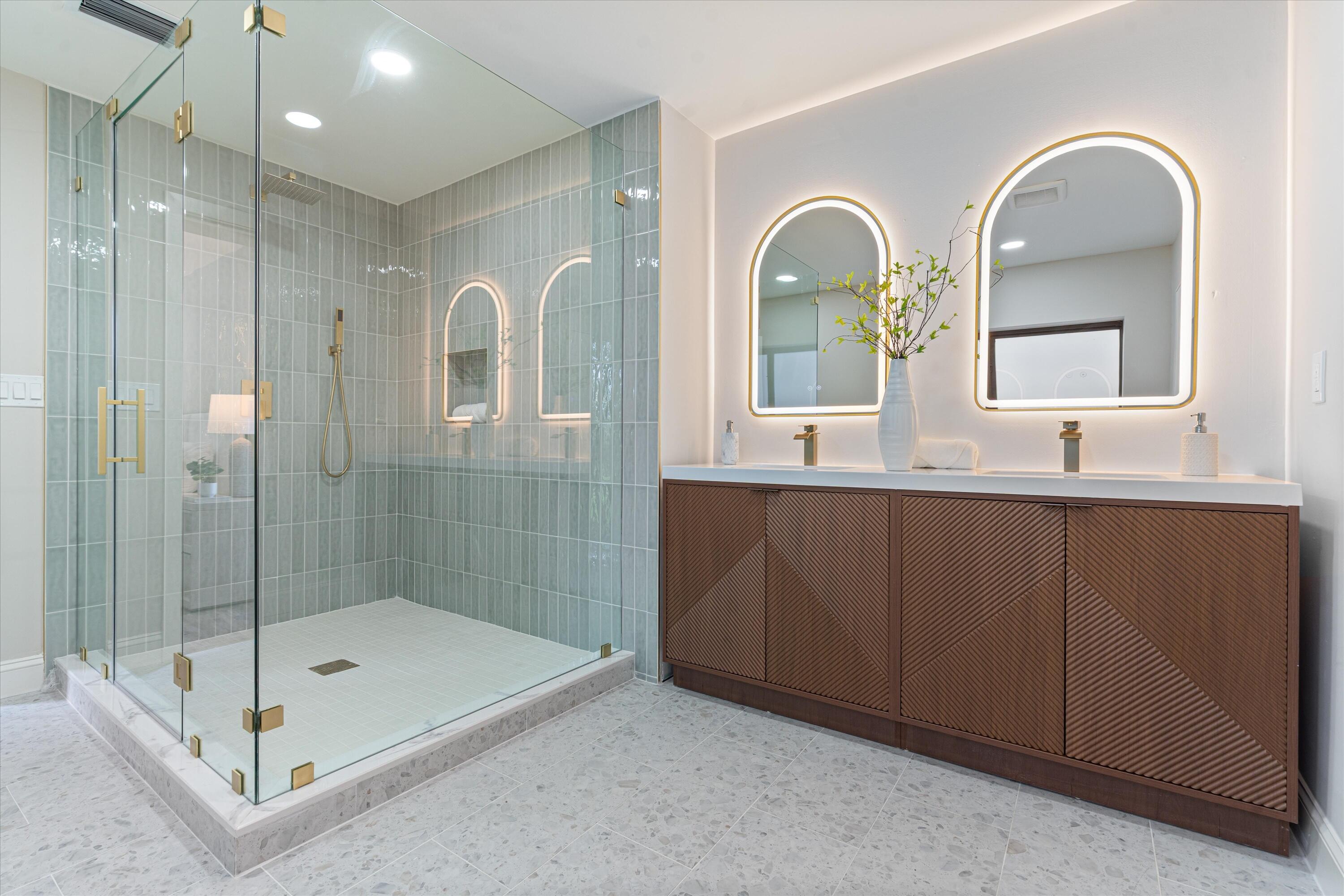 Double vanity featuring textured wood cabinetry, stone countertops, integrated arched LED-backlit mirrors, gold-finish hardware, and a glass-enclosed shower with vertical wall tile