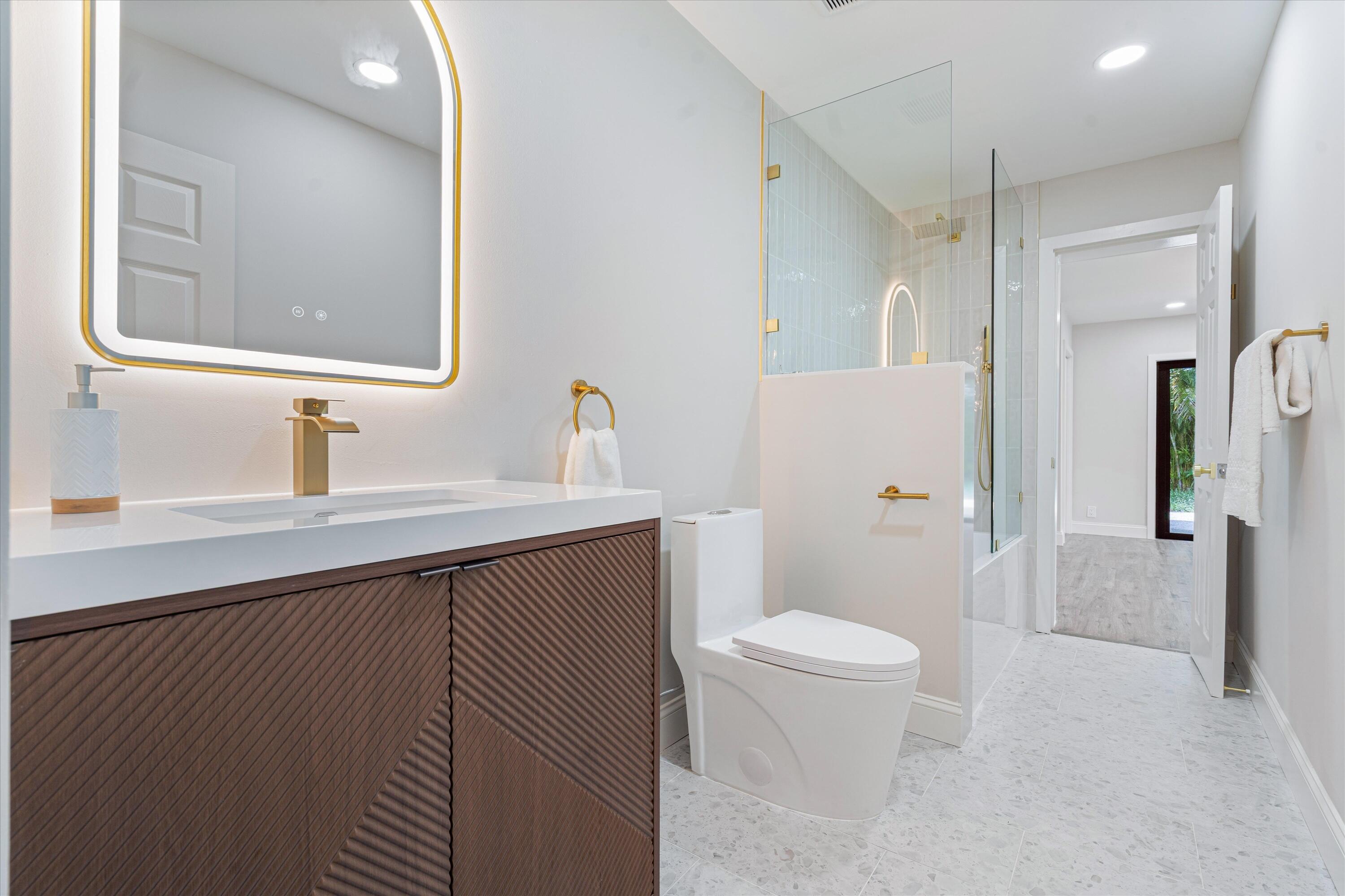 Floating vanity with a textured wood finish, solid surface countertop, brass-toned hardware, arched LED-backlit mirror, and glass-enclosed shower