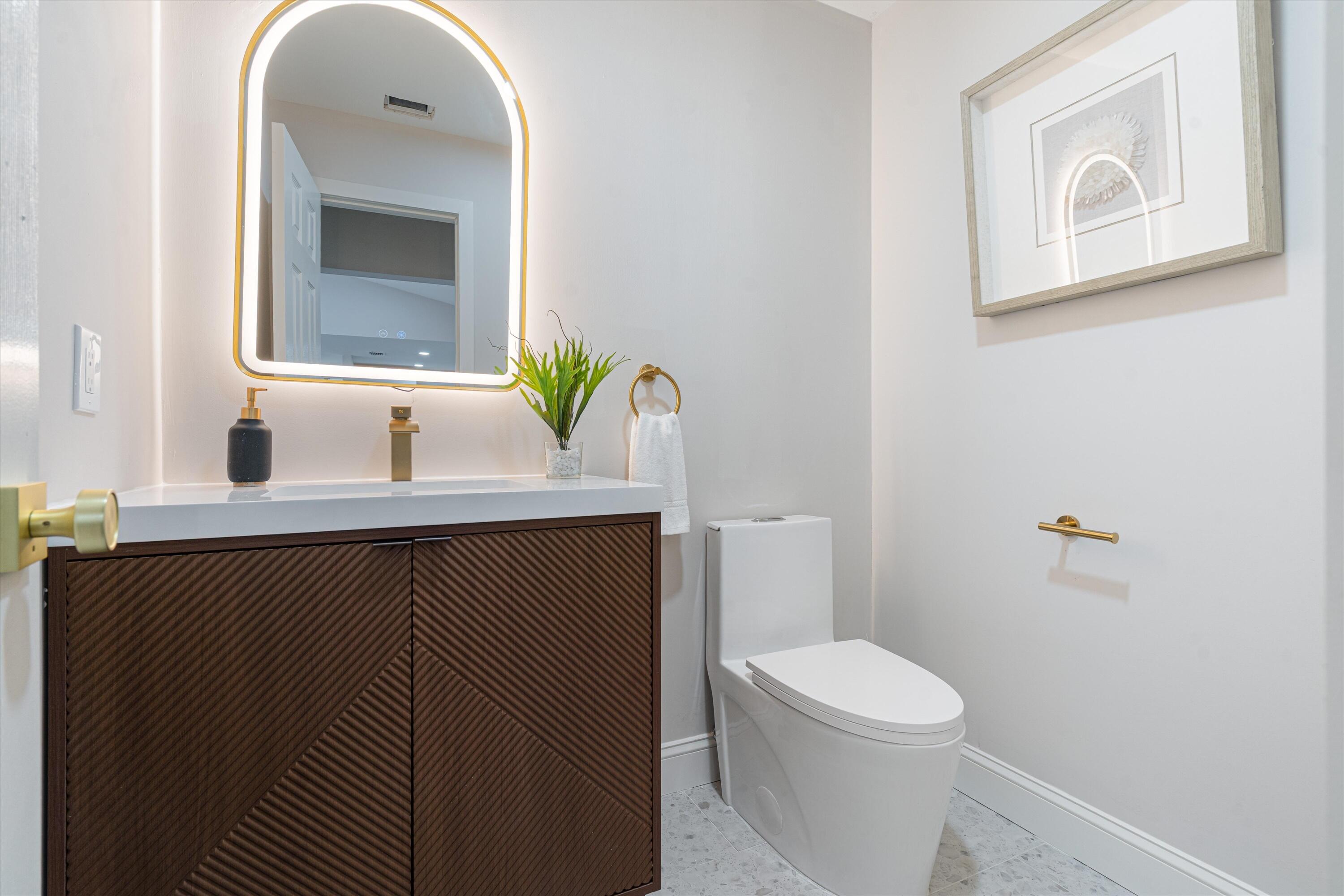 Modern vanity with textured wood-finish cabinet doors, white surface, arched backlit mirror, brass-finish fixtures, and a one-piece dual-flush toilet