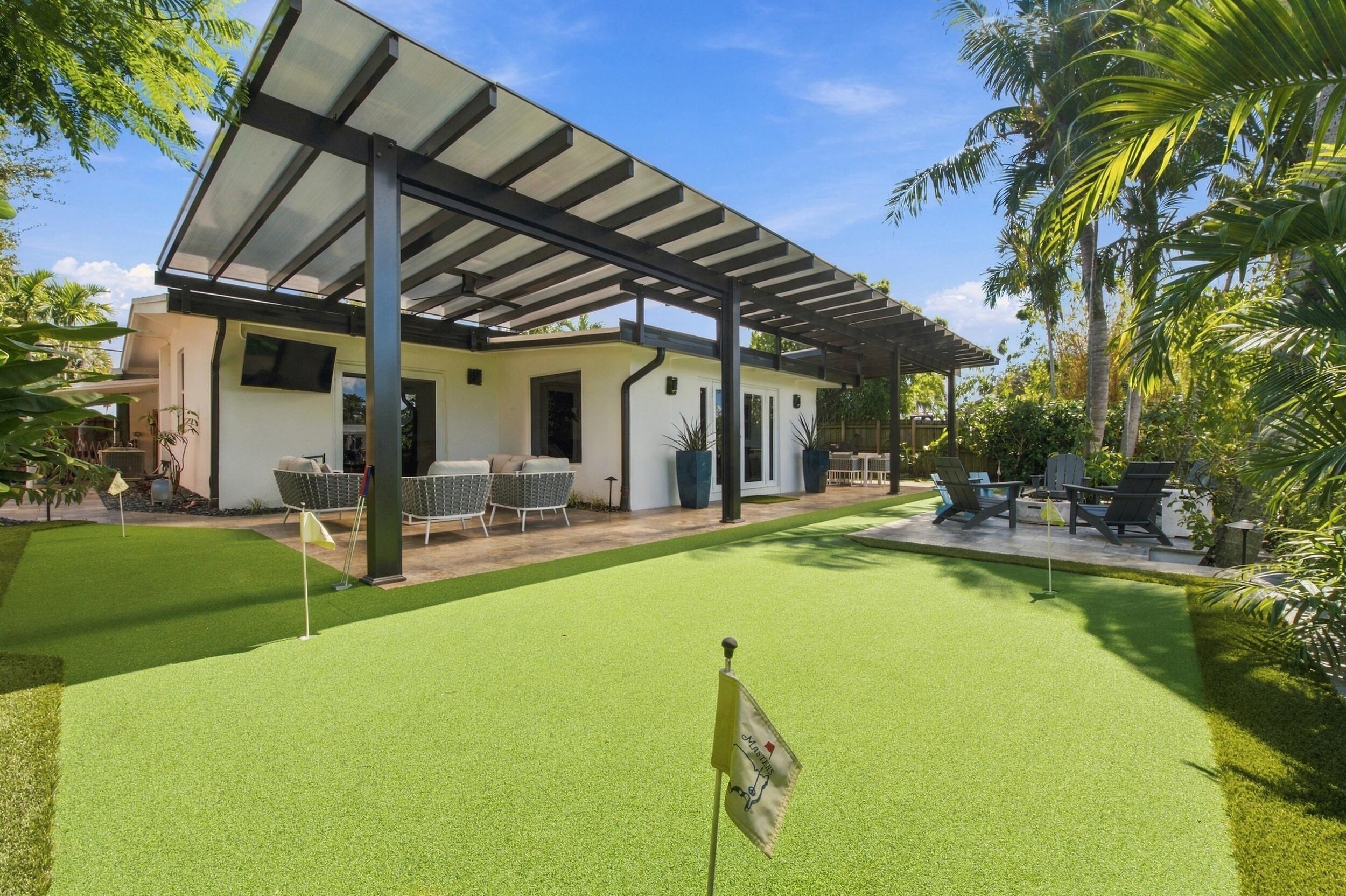 Backyard featuring a synthetic turf putting green with multiple holes, covered patio with black metal structural framing, and hardscaped stone flooring