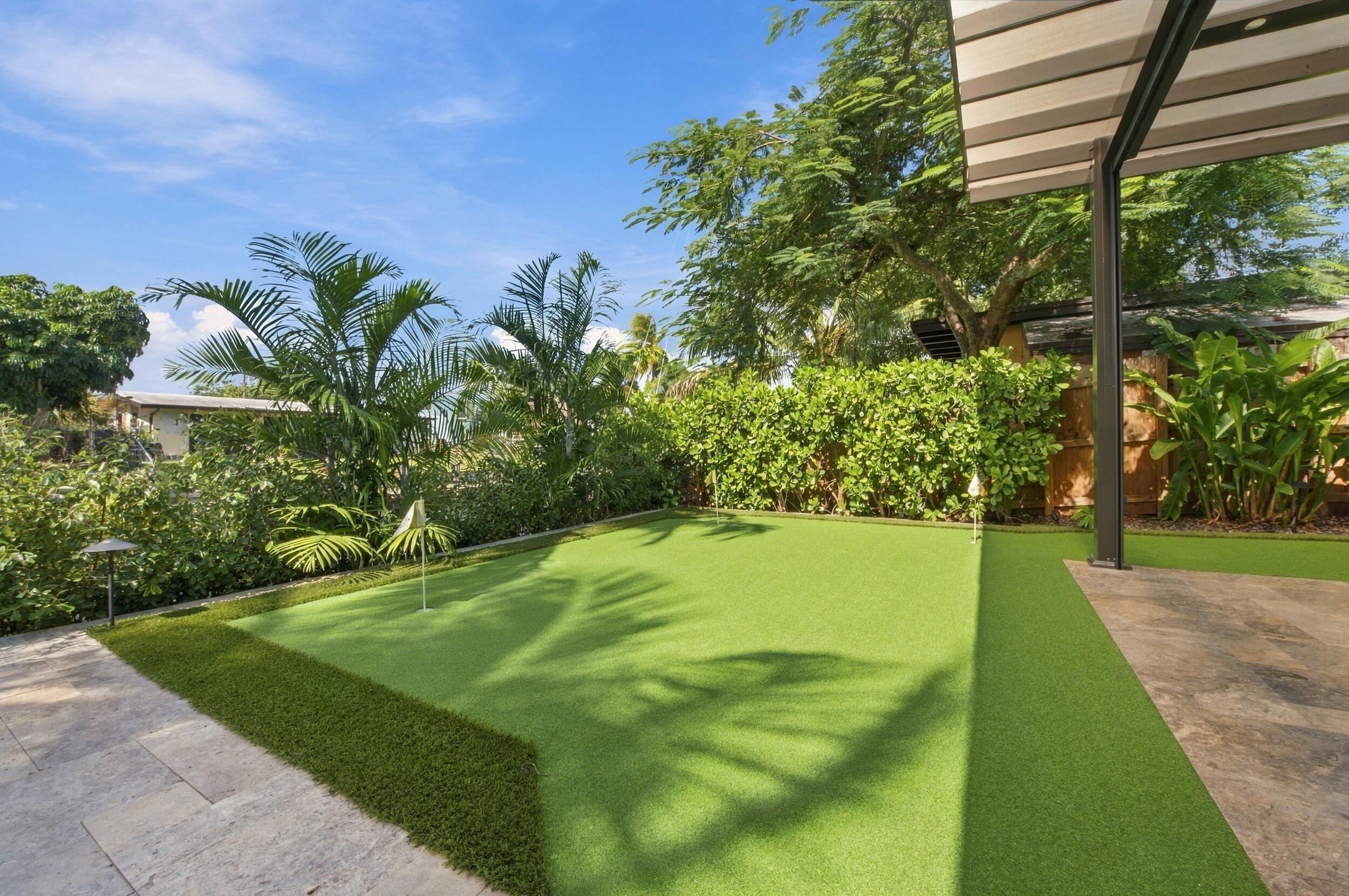 Backyard putting green featuring synthetic turf, stone-paver patio, metal structural awning, and privacy hedging