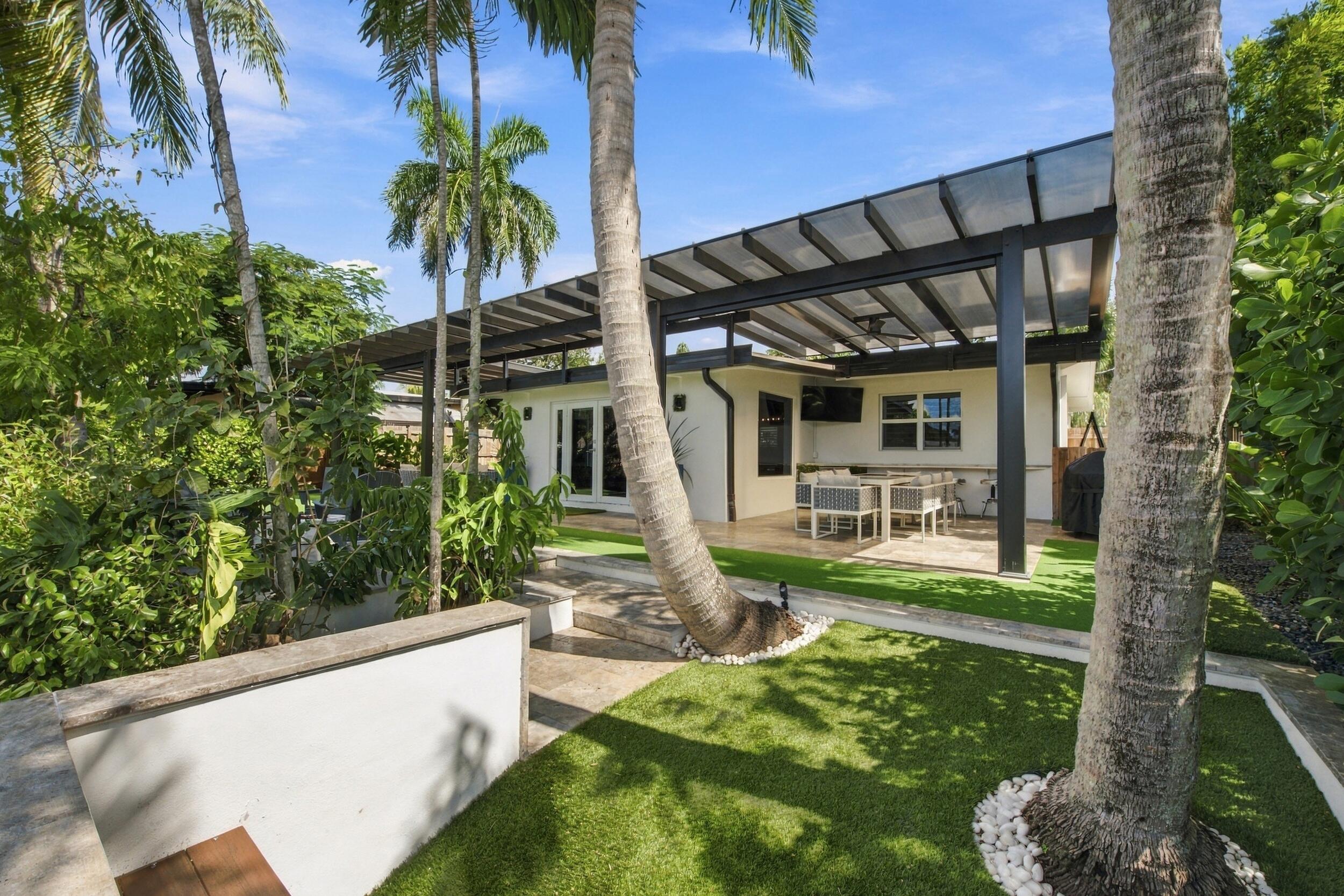 Outdoor living space featuring a modern pergola with a semi-translucent roof, stone patio flooring, synthetic lawn, and mature palm trees