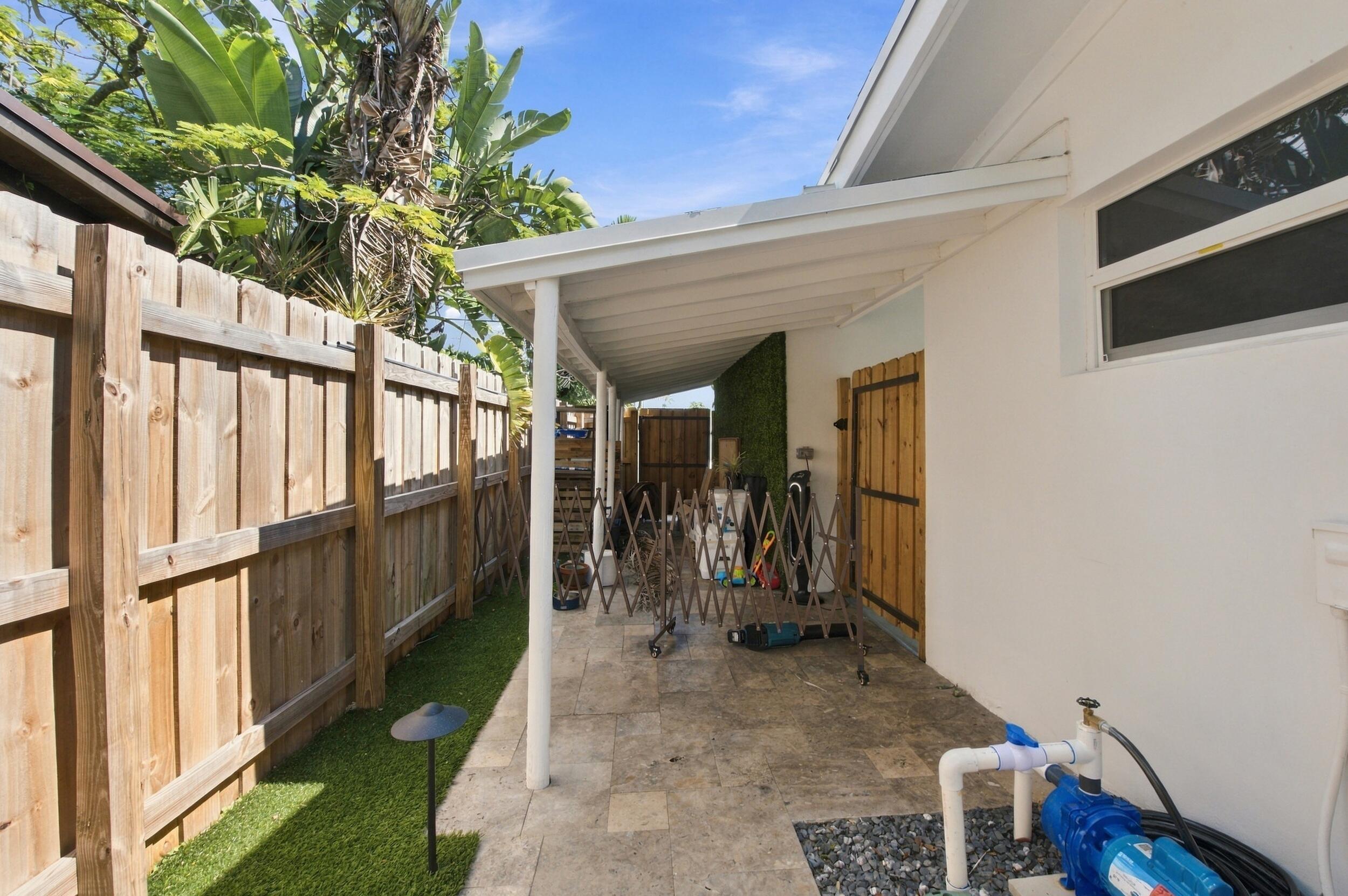 Covered patio featuring stone tile flooring, enclosed by a wood privacy fence, synthetic turf lawn, and a custom wood side gate