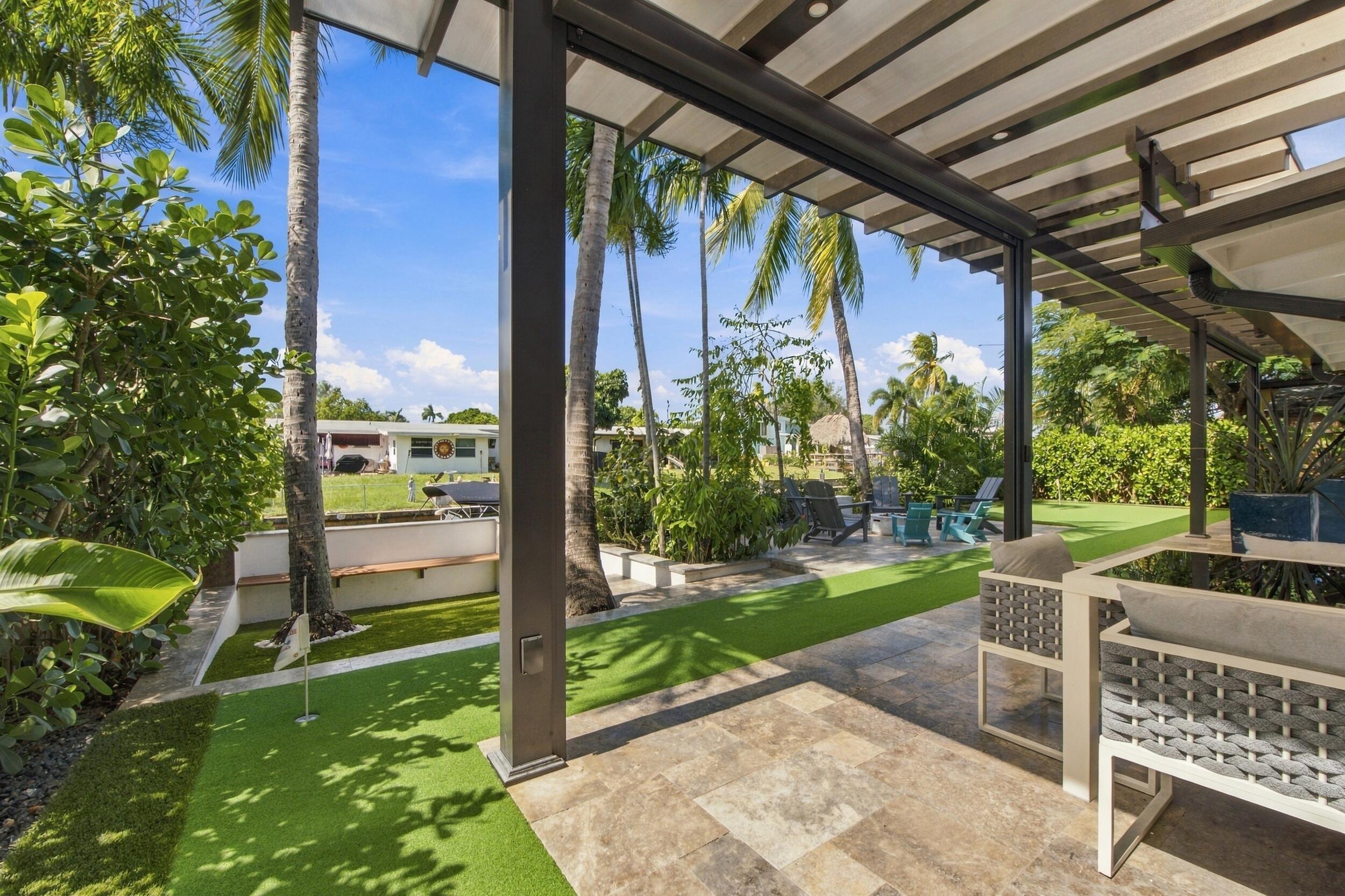 Covered patio featuring stone-tile flooring, louvered overhead structure, synthetic turf yard, built-in masonry seating, and mature palm trees