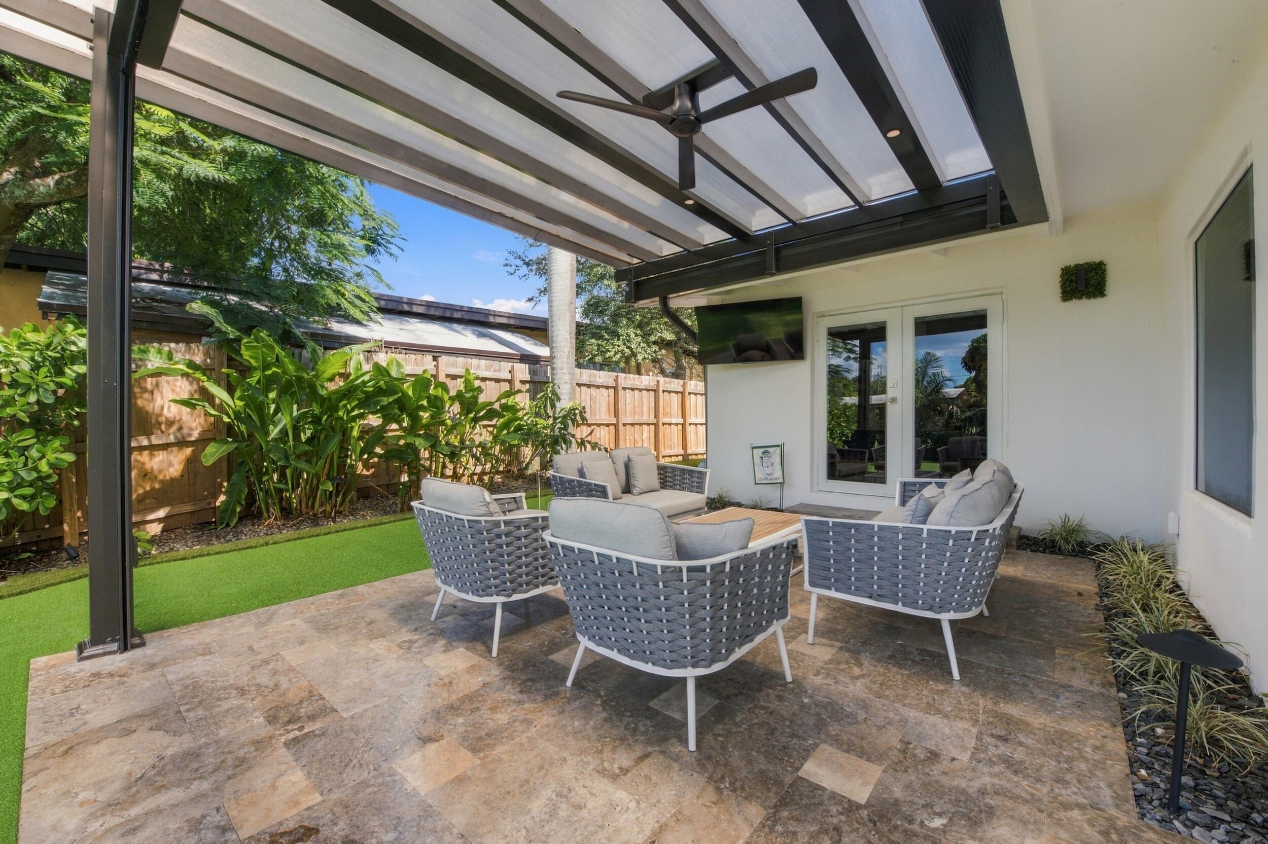 Covered outdoor patio featuring large-format stone tiling, a fixed overhead pergola, a built-in ceiling fan, and synthetic turf lawn