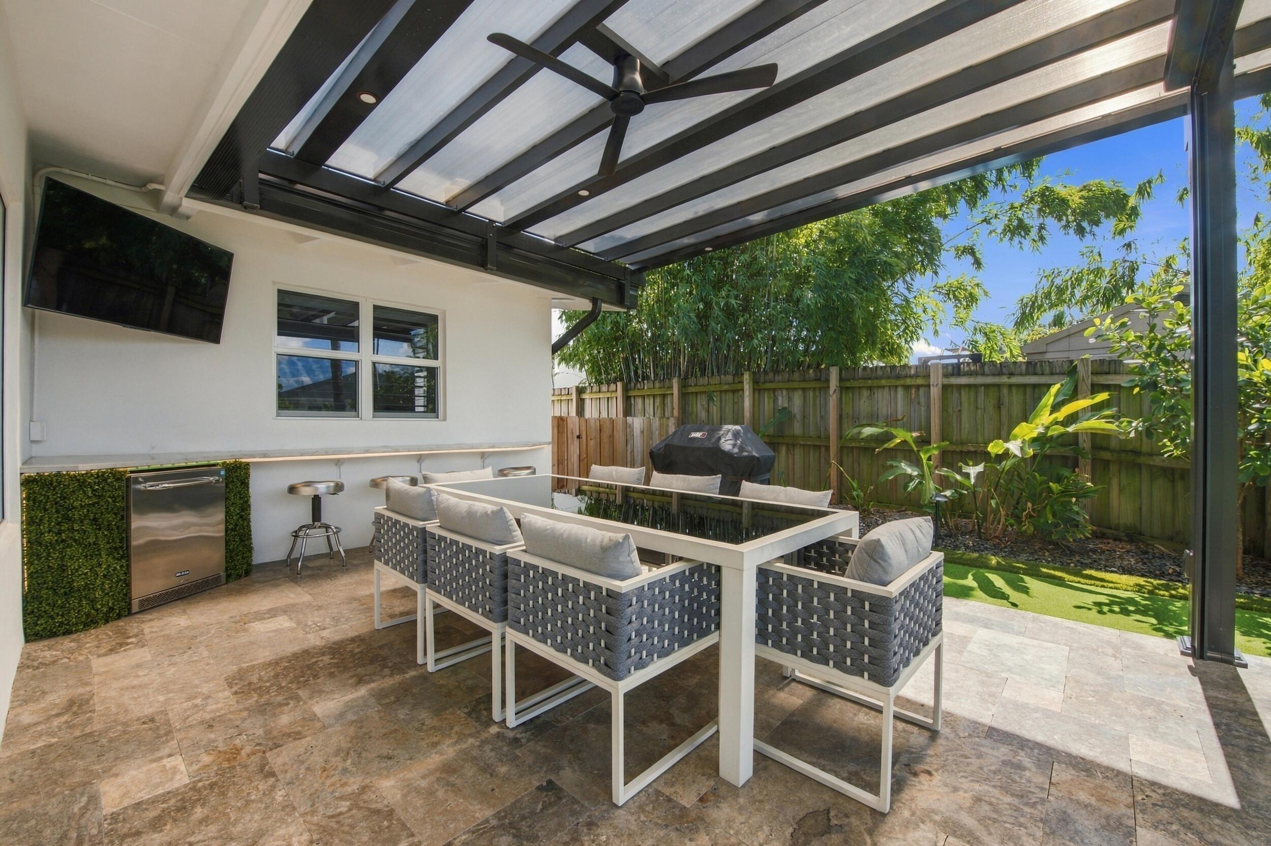 Covered patio featuring stone tile flooring, a built-in outdoor kitchen counter with an under-counter refrigerator, a black metal pergola, and a ceiling fan