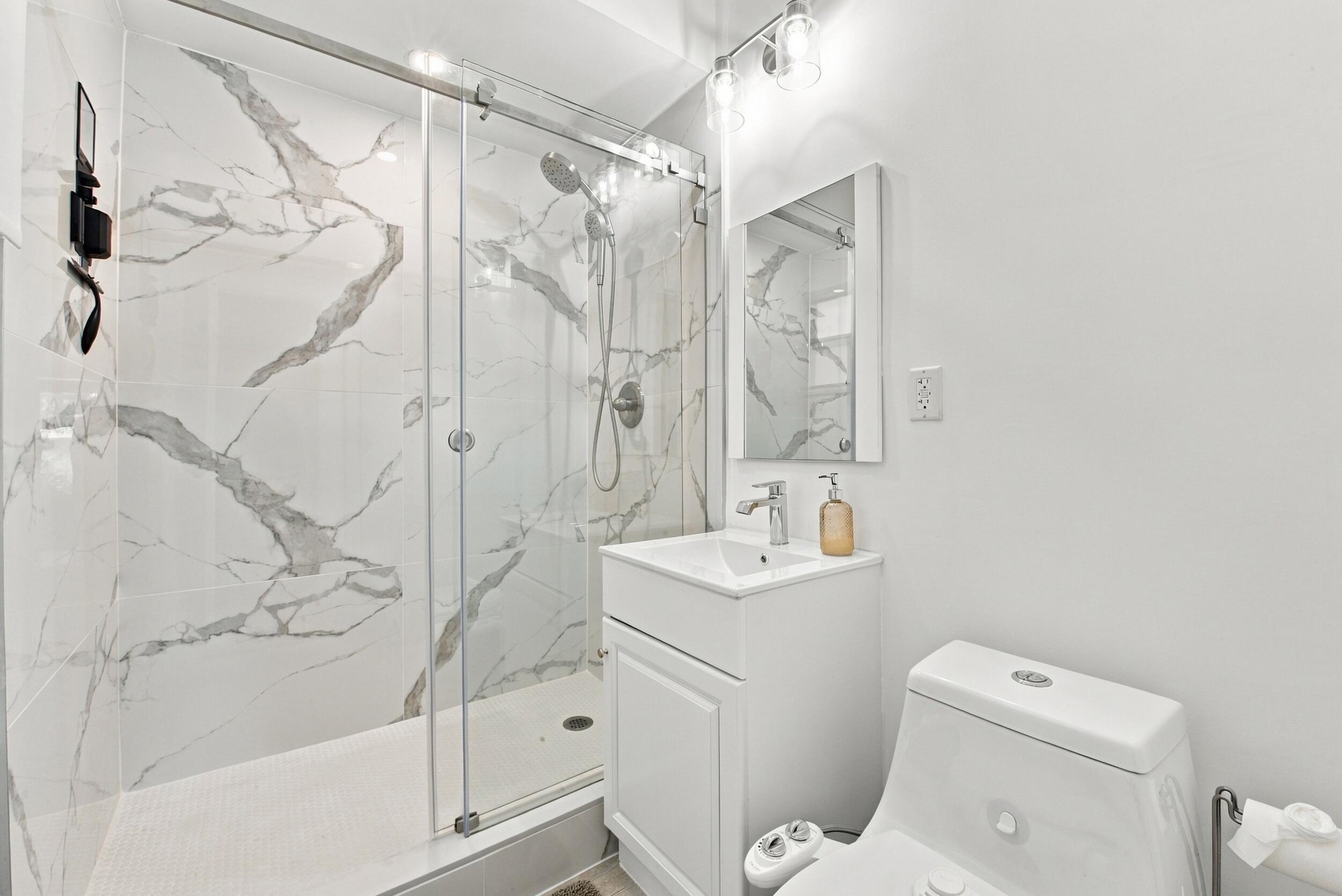 Walk-in shower with floor-to-ceiling stone-patterned tile, glass sliding enclosure, handheld shower fixture, white vanity with rectangular sink, and polished chrome hardware