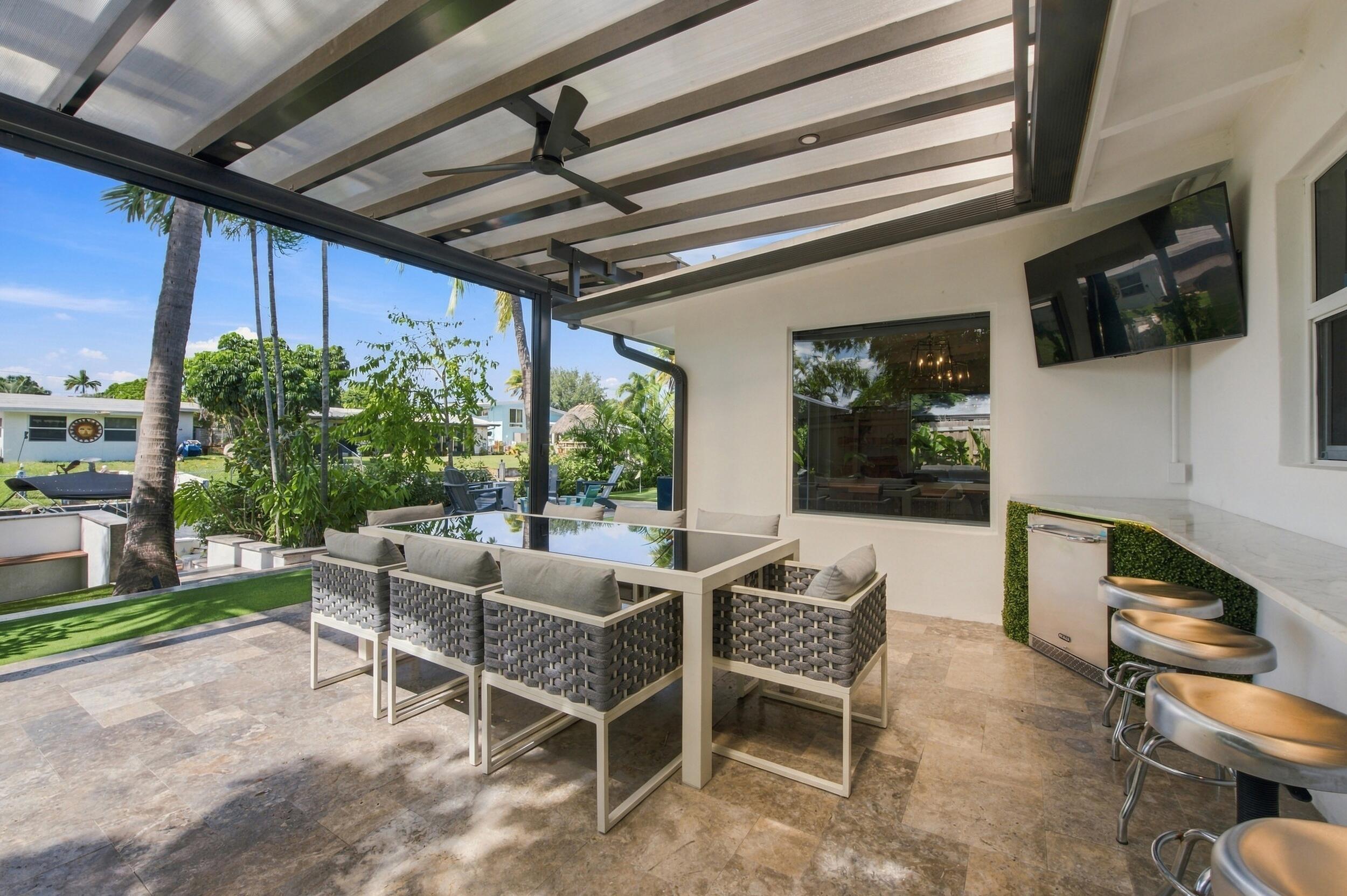Covered patio featuring stone-tiled flooring, a slatted timber pergola, a built-in outdoor bar with a stone countertop, an integrated ceiling fan, and a mounted flat-screen television