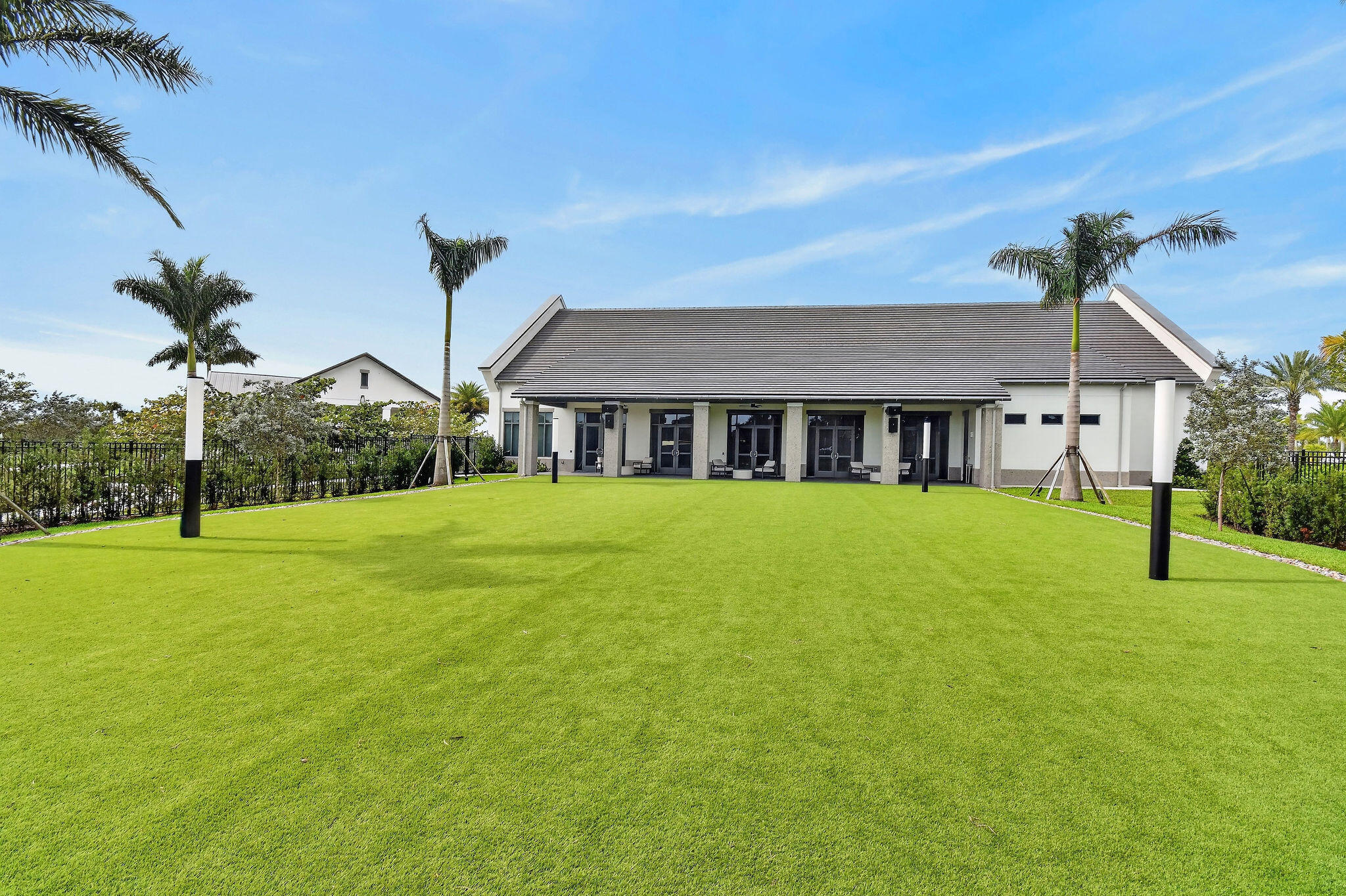 Expansive green lawn with established palm trees, perimeter fencing, and a modern building with a dark-tiled roof and multiple glass doors