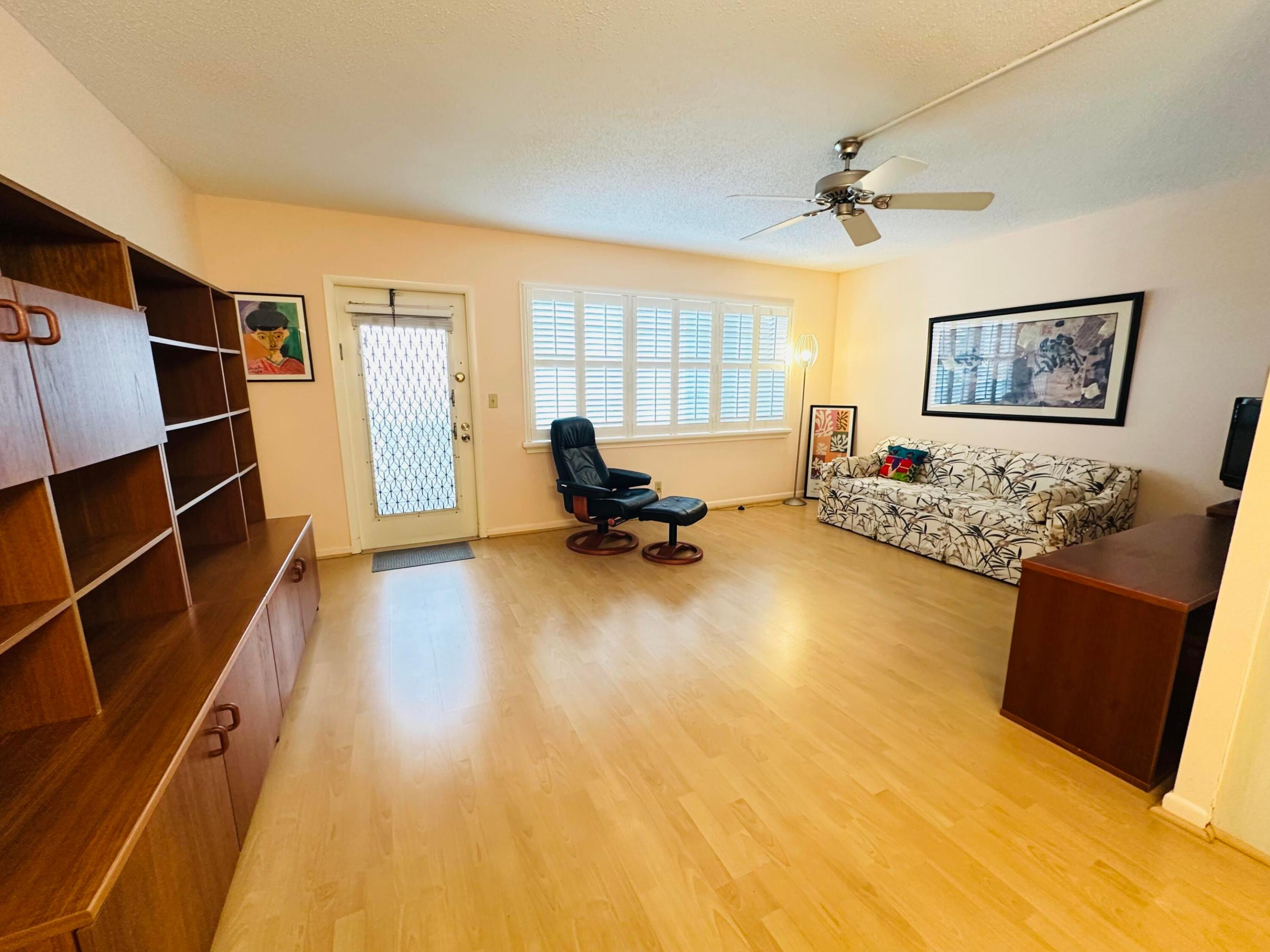 Living area featuring laminate flooring, wall unit, multi-panel window with white plantation-style shutters, and a ceiling fan Living area featuring laminate flooring, wall unit, multi-panel window with white plantation-style shutters, and a ceiling fan