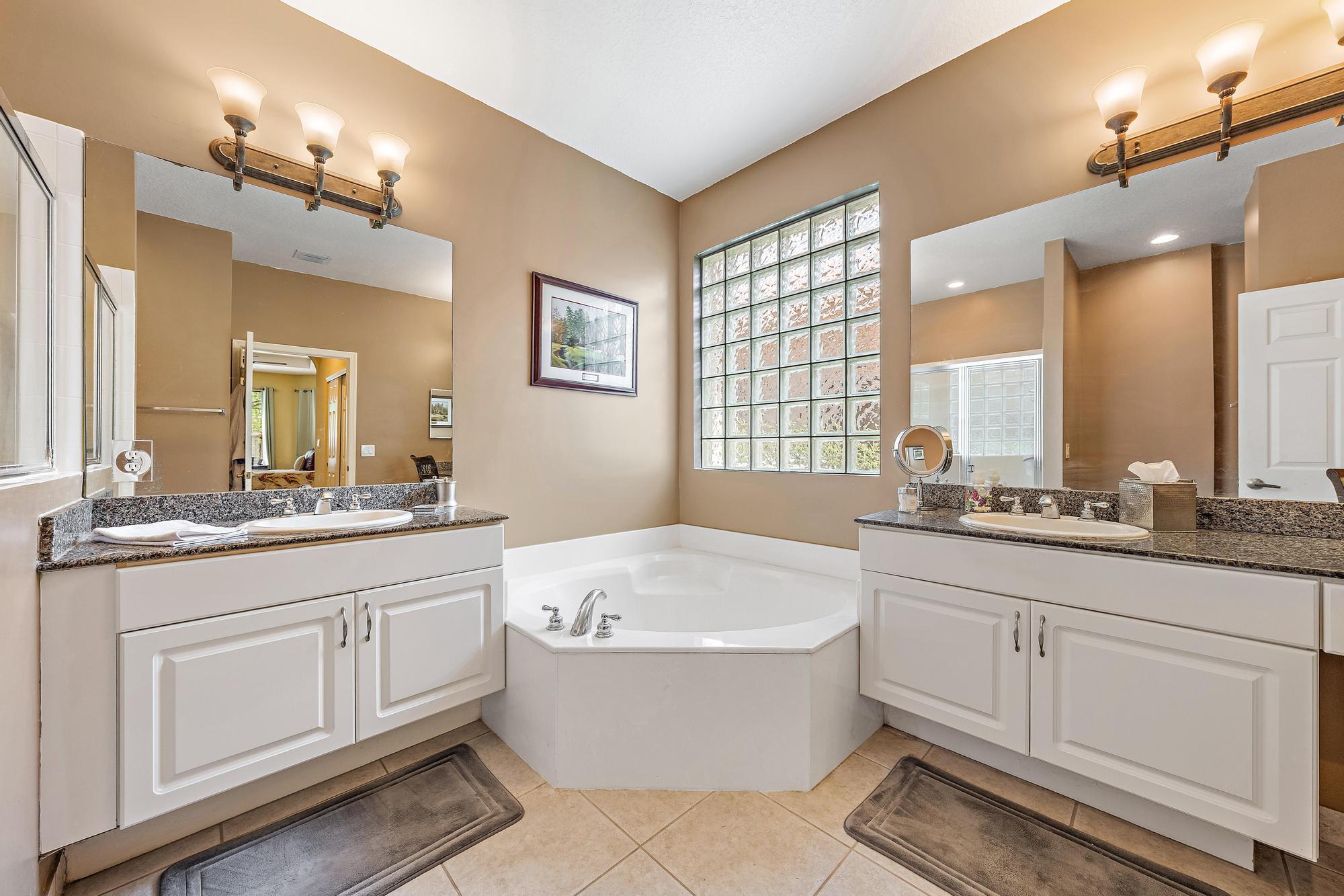 Dual white vanity cabinets featuring stone-surface countertops, integrated oval sinks, glass block accent window, and a built-in corner soaking tub