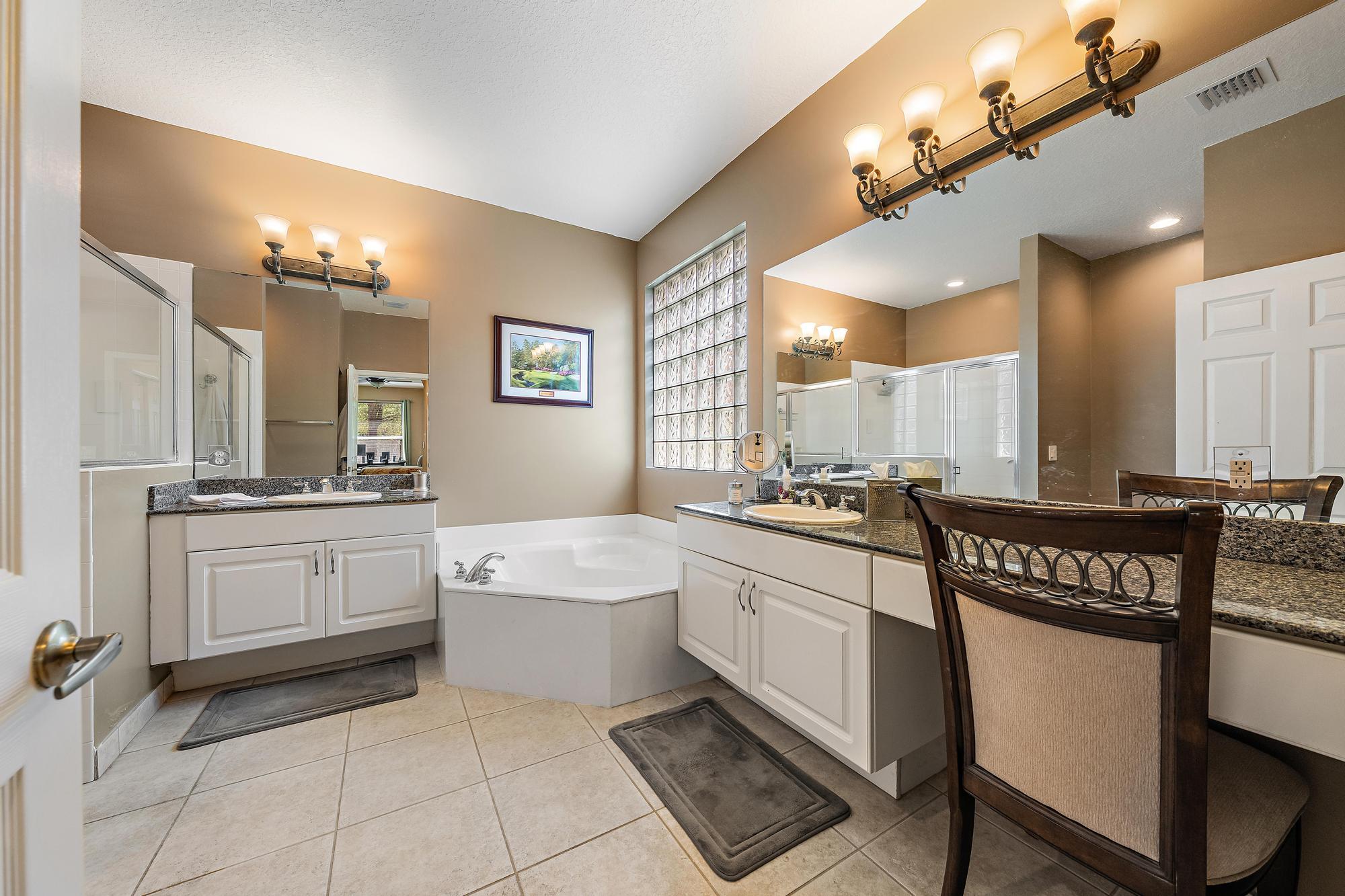Bathroom featuring dual white cabinetry with stone-surface countertops, soaking tub, glass-block window, and ceramic tile flooring