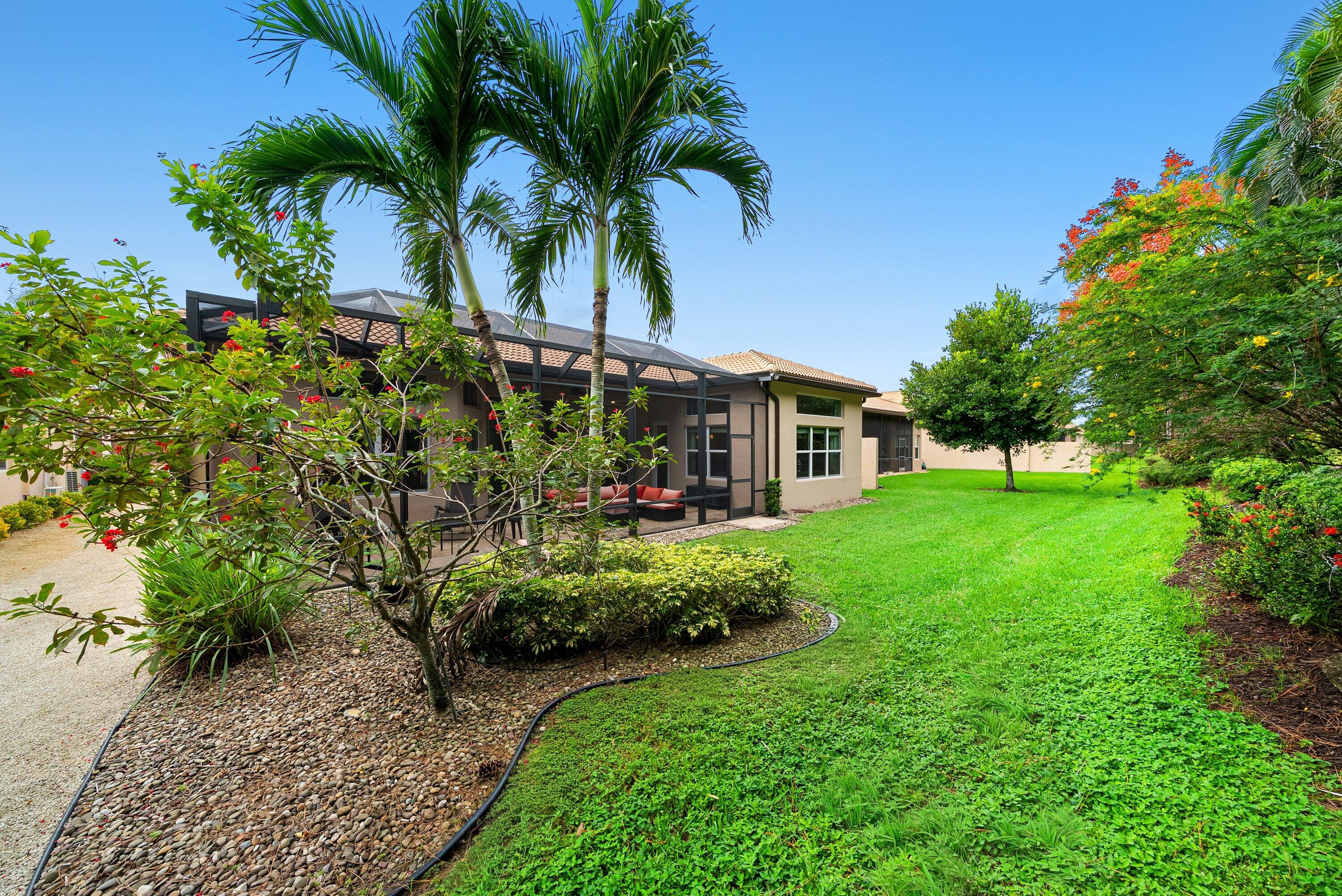 Backyard featuring a screened-in lanai, manicured lawn, established palm trees, decorative rock landscaping, and perimeter shrubbery