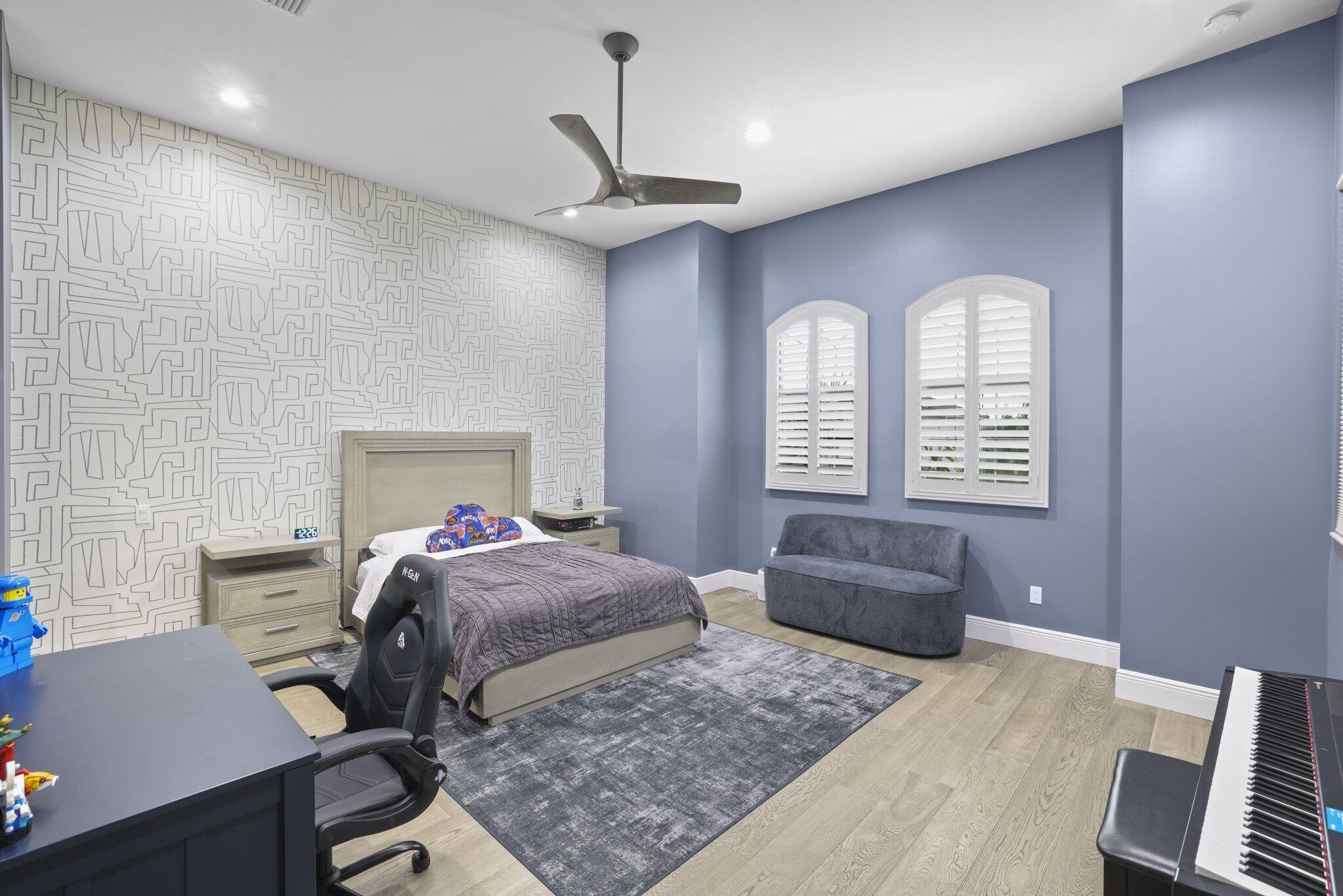Light wood-finish flooring, accent wall with patterned wallpaper, arched windows with plantation shutters, ceiling fan, and recessed lighting