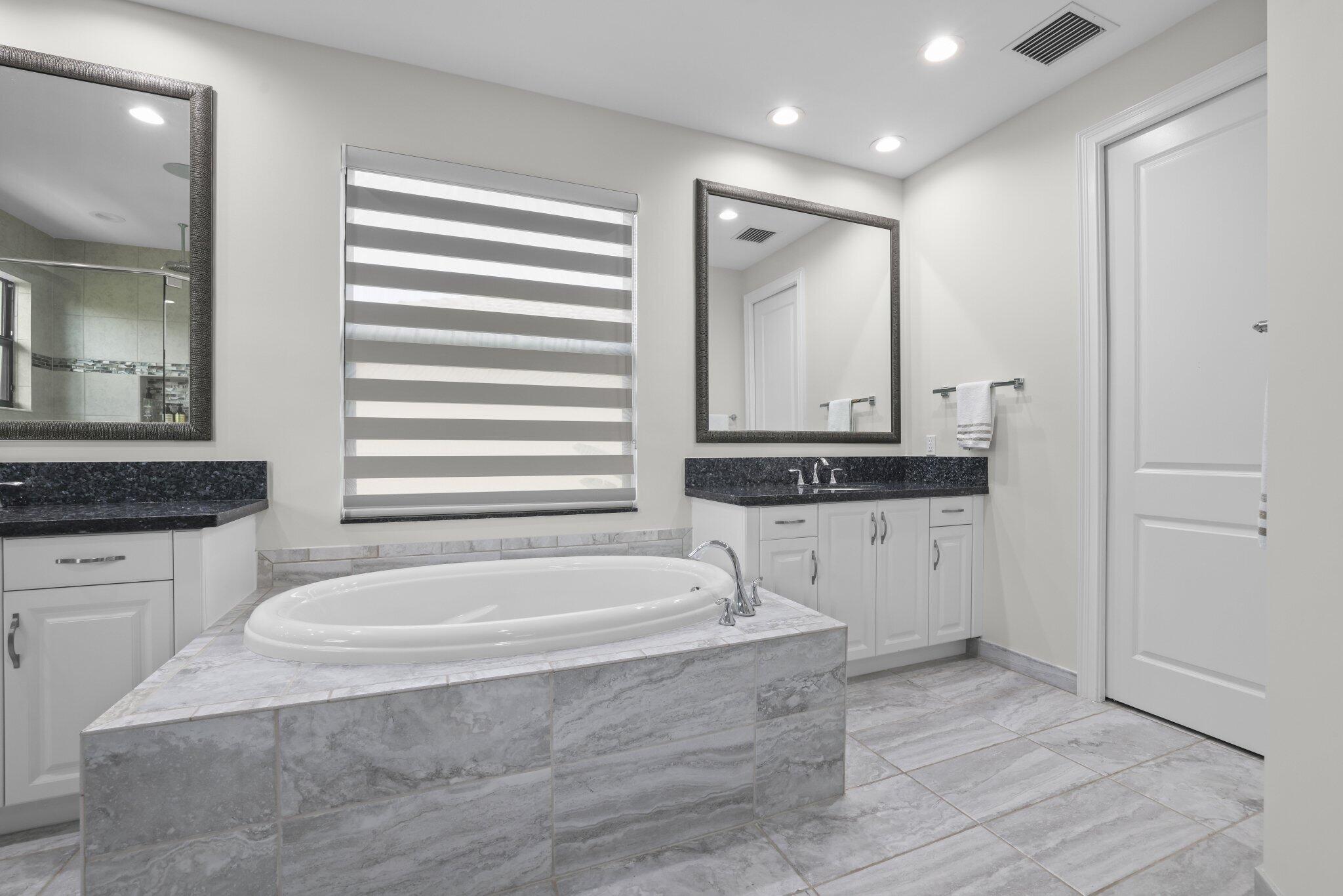 Built-in soaking tub with a tiled surround, dual vanities with dark countertops, and tiled flooring