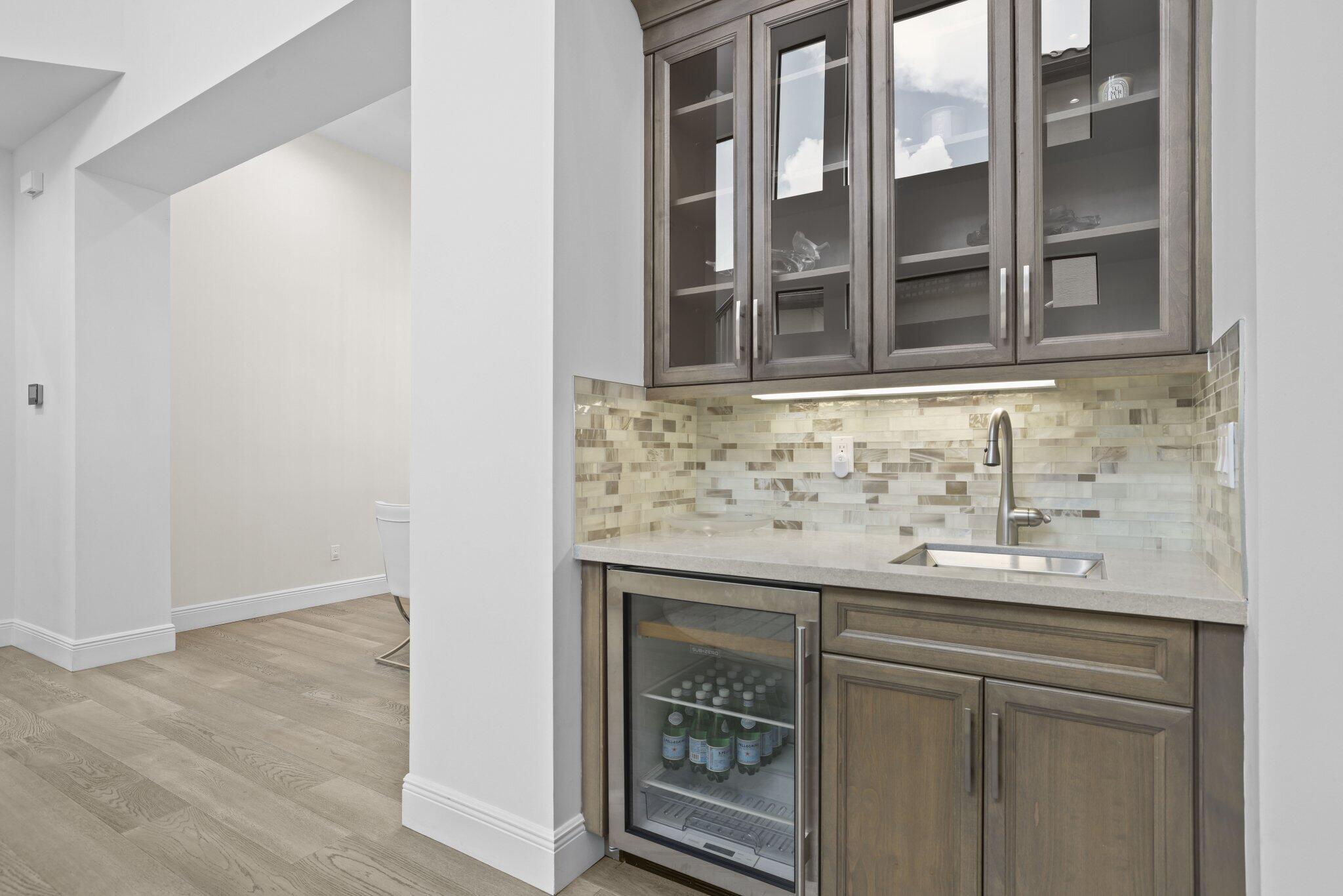 Built-in wet bar featuring gray cabinetry, glass-front upper cabinets, light-toned countertop, mosaic tile backsplash, and a beverage refrigerator