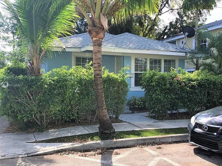 Exterior featuring light blue lap siding, multi-pane windows with white trim, and a concrete walkway bordered by mature privacy hedges and a palm tree Exterior featuring light blue lap siding, multi-pane windows with white trim, and a concrete walkway bordered by mature privacy hedges and a palm tree