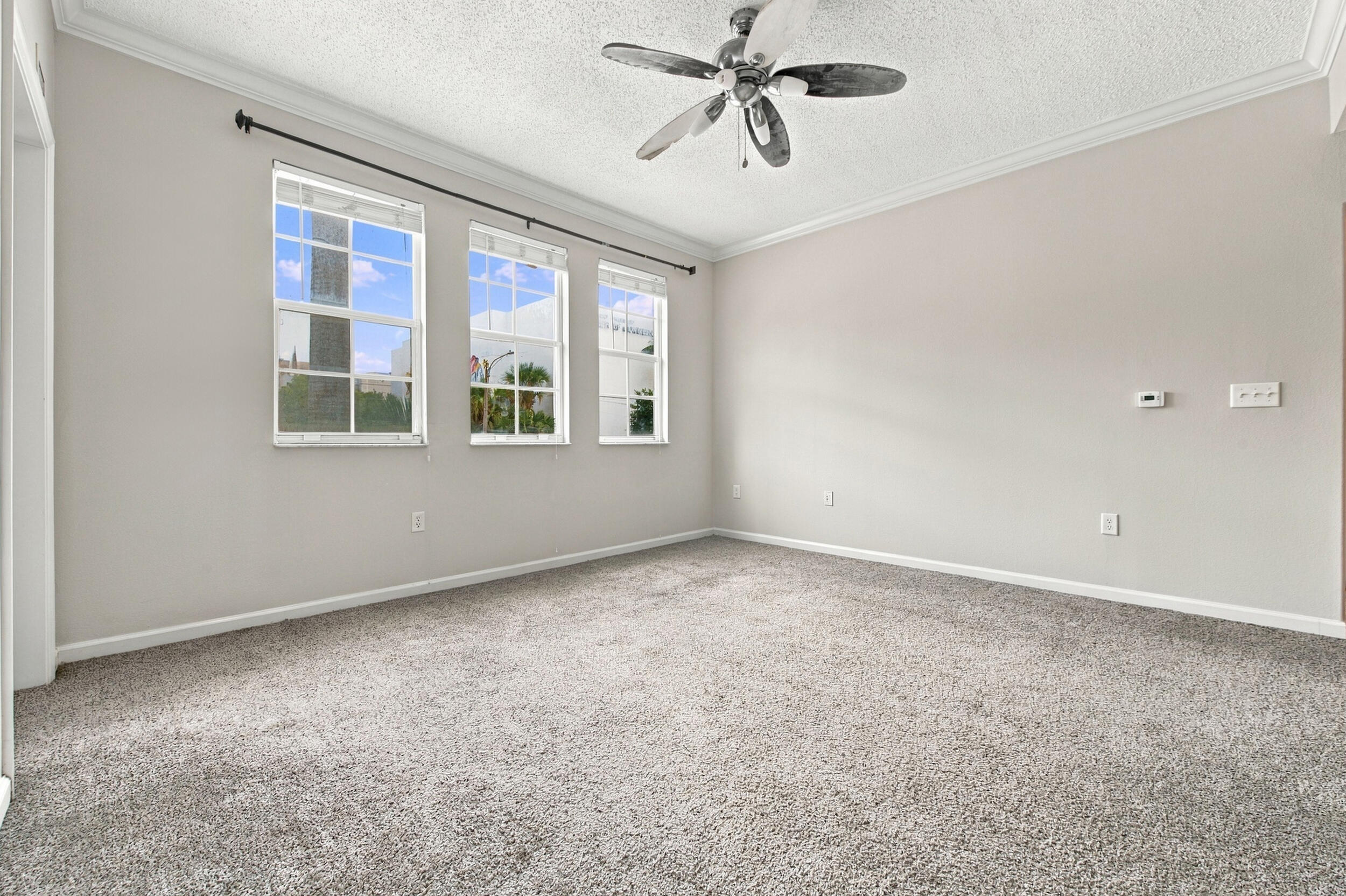 Neutral-toned interior featuring wall-to-wall carpeting, three multi-pane windows, crown molding, and a ceiling fan