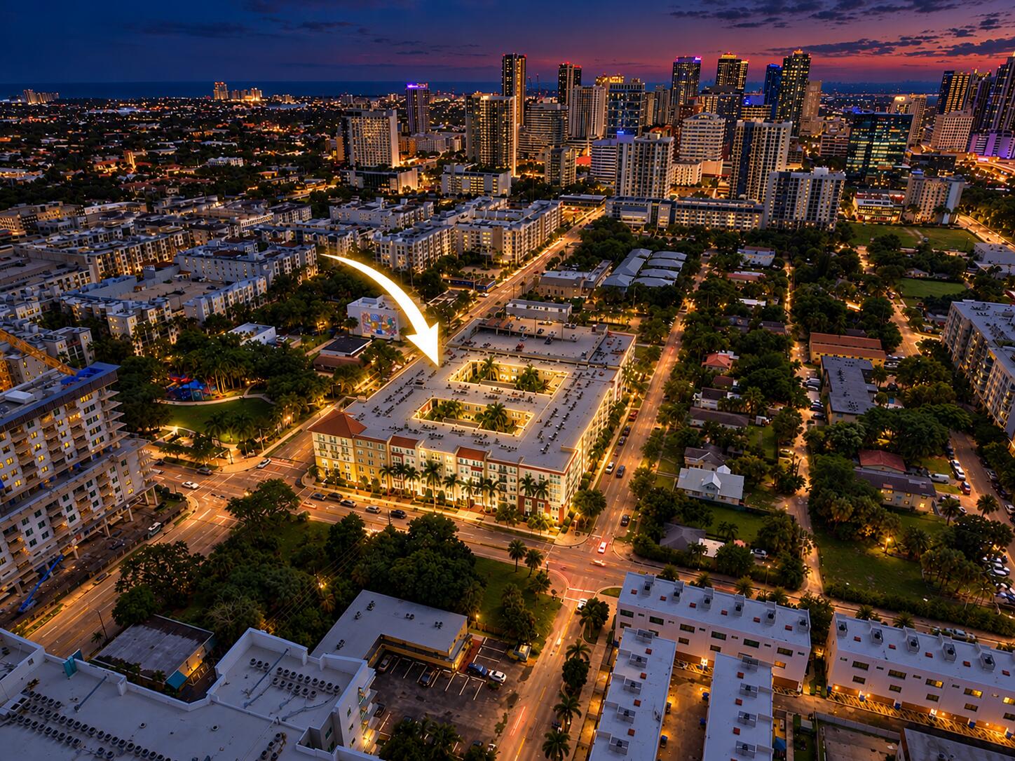 Aerial perspective of a multi-story residential building featuring a central courtyard with mature palms, a tile roof, and urban skyline horizon