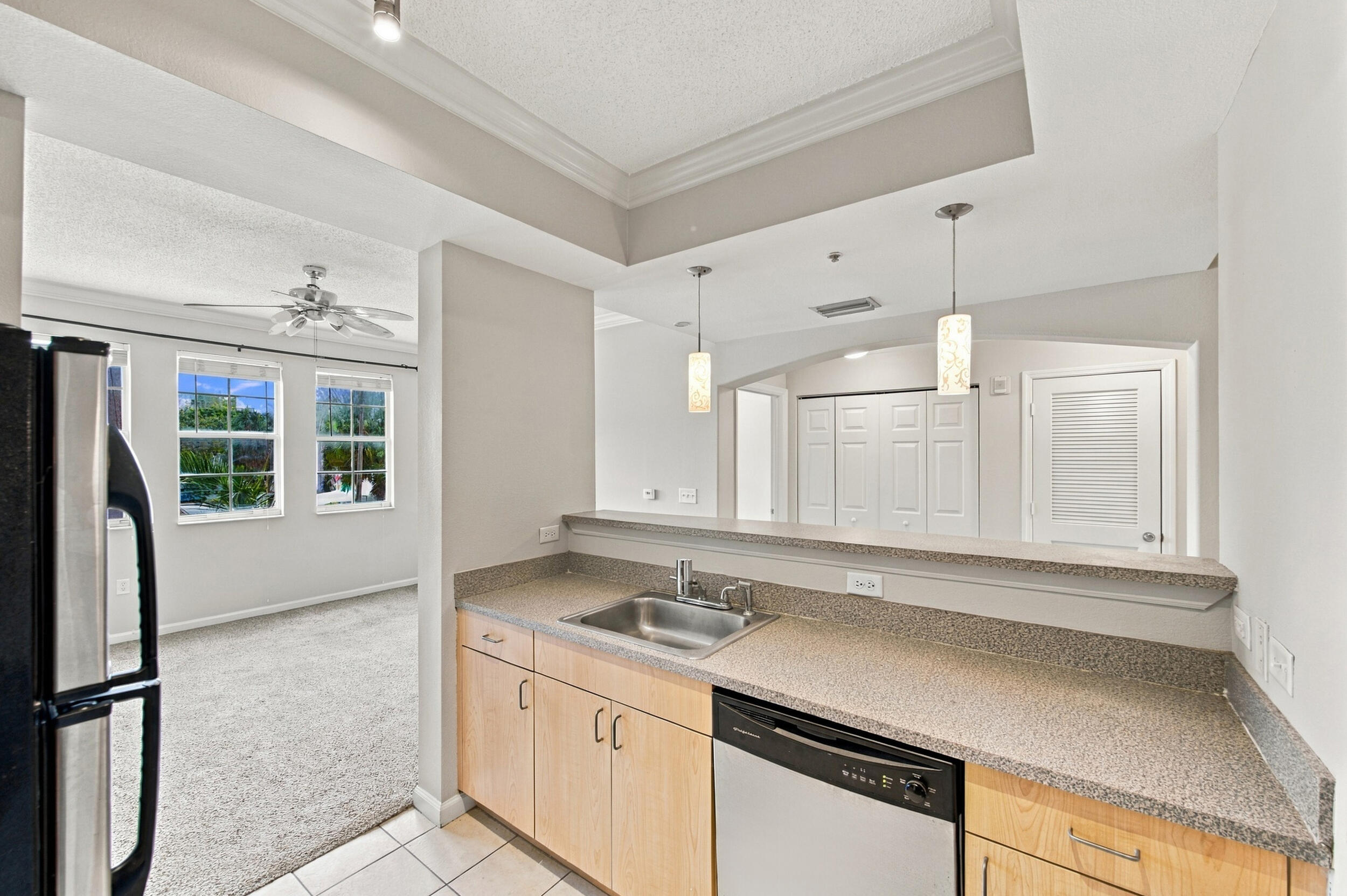 Kitchen featuring light wood-tone cabinetry, solid surface countertops, stainless steel undermount sink, and integrated stainless steel dishwasher, illuminated by suspended pendant lighting